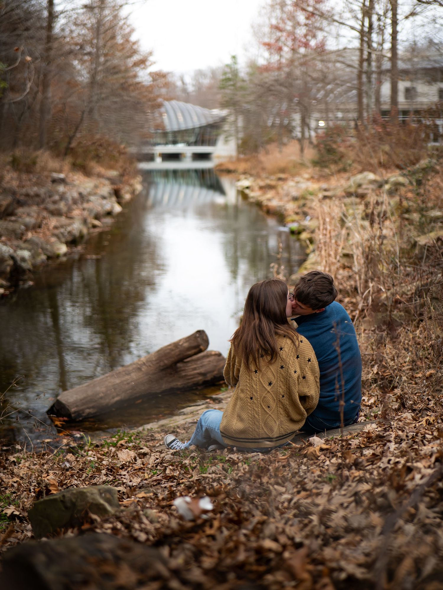 Two people sit together on fallen leaves by a tranquil stream on an autumn afternoon.