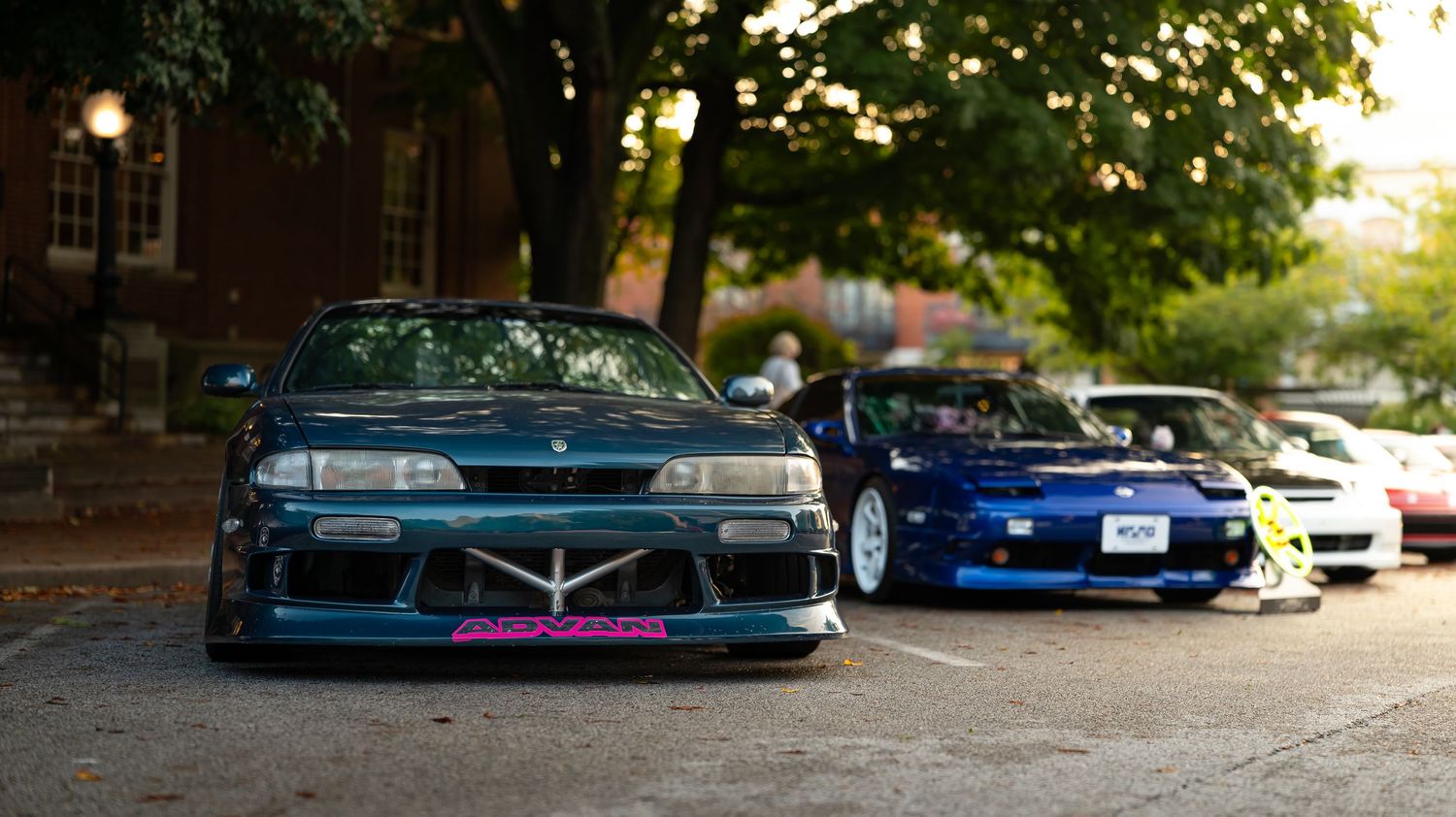 Modified performance vehicles lined up at a car show with brick buildings visible.