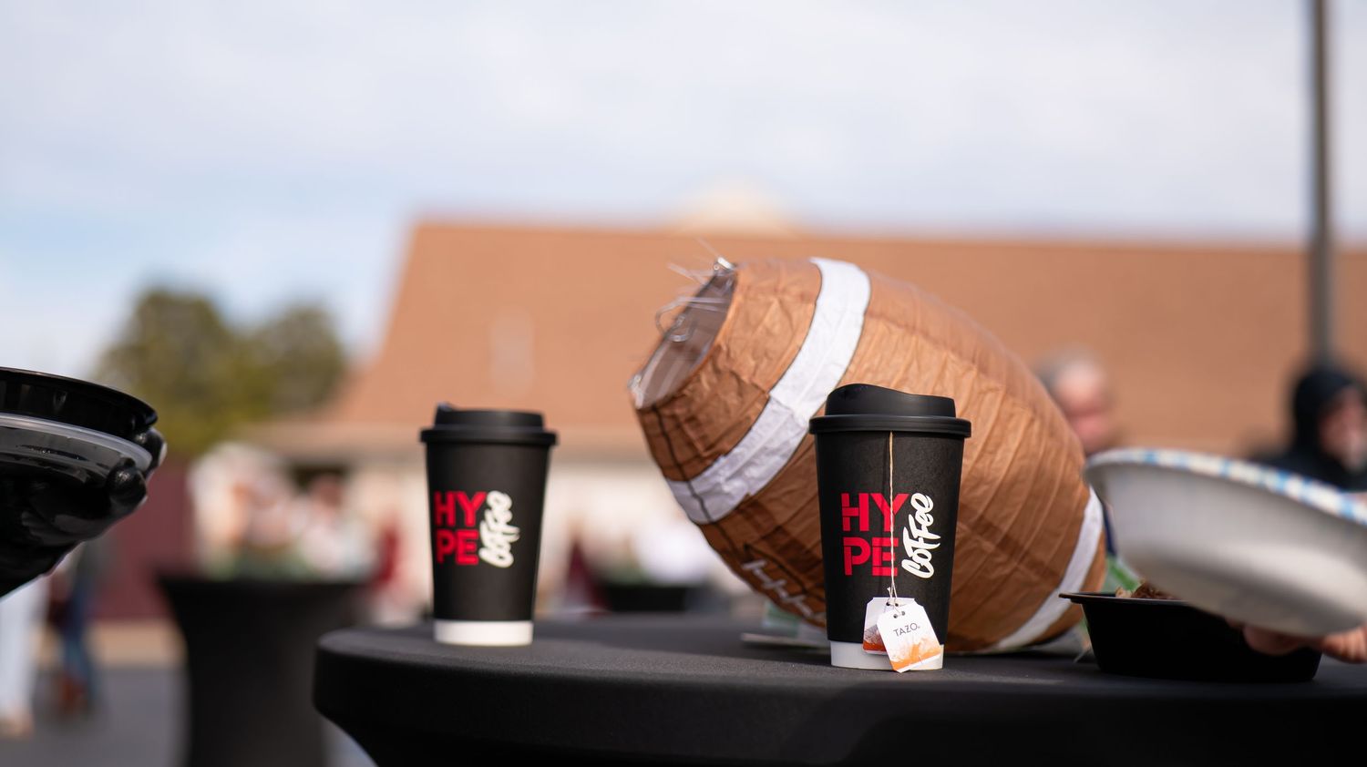 Coffee cups and snacks arranged on a dark surface at an outdoor event.