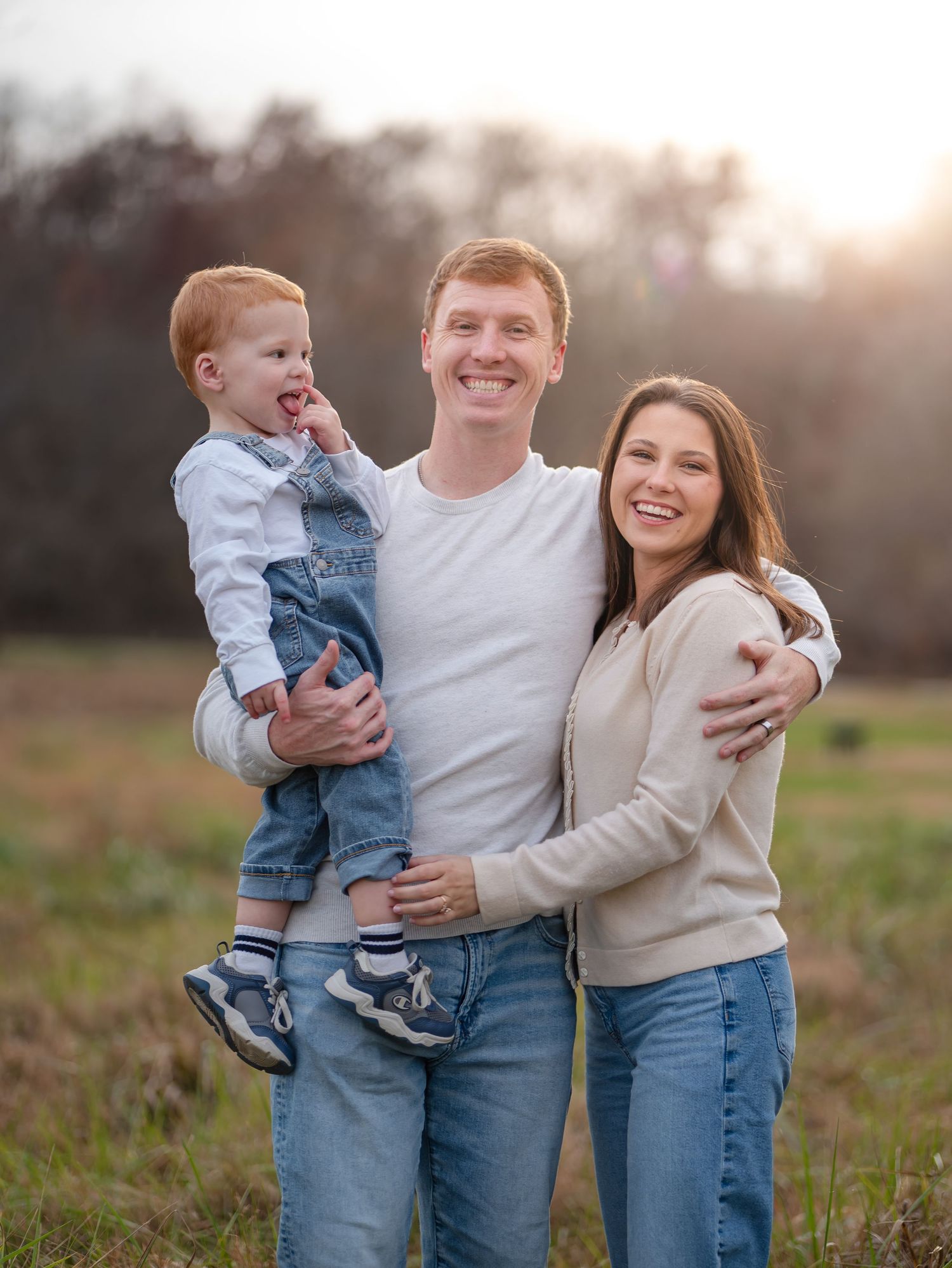 A family of three wearing casual clothing poses together in a sunlit field during golden hour.