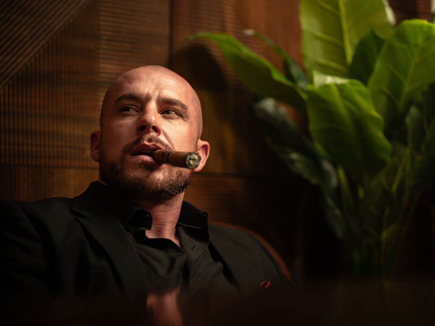 Bald man smoking a cigar in a dimly lit room with tropical plants in the background.