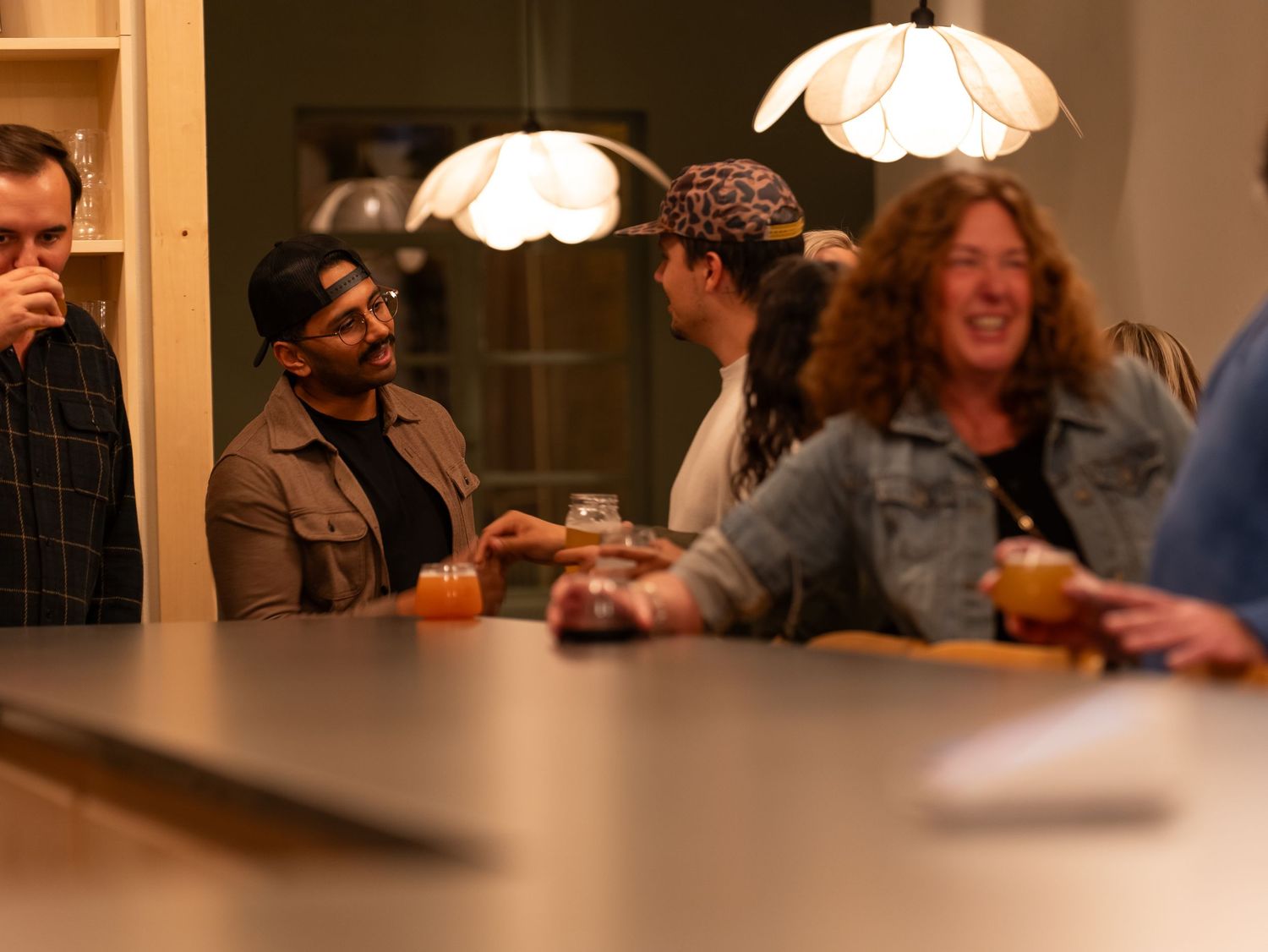 People gathered around a table enjoying drinks under pendant lamps in a warm, social setting.