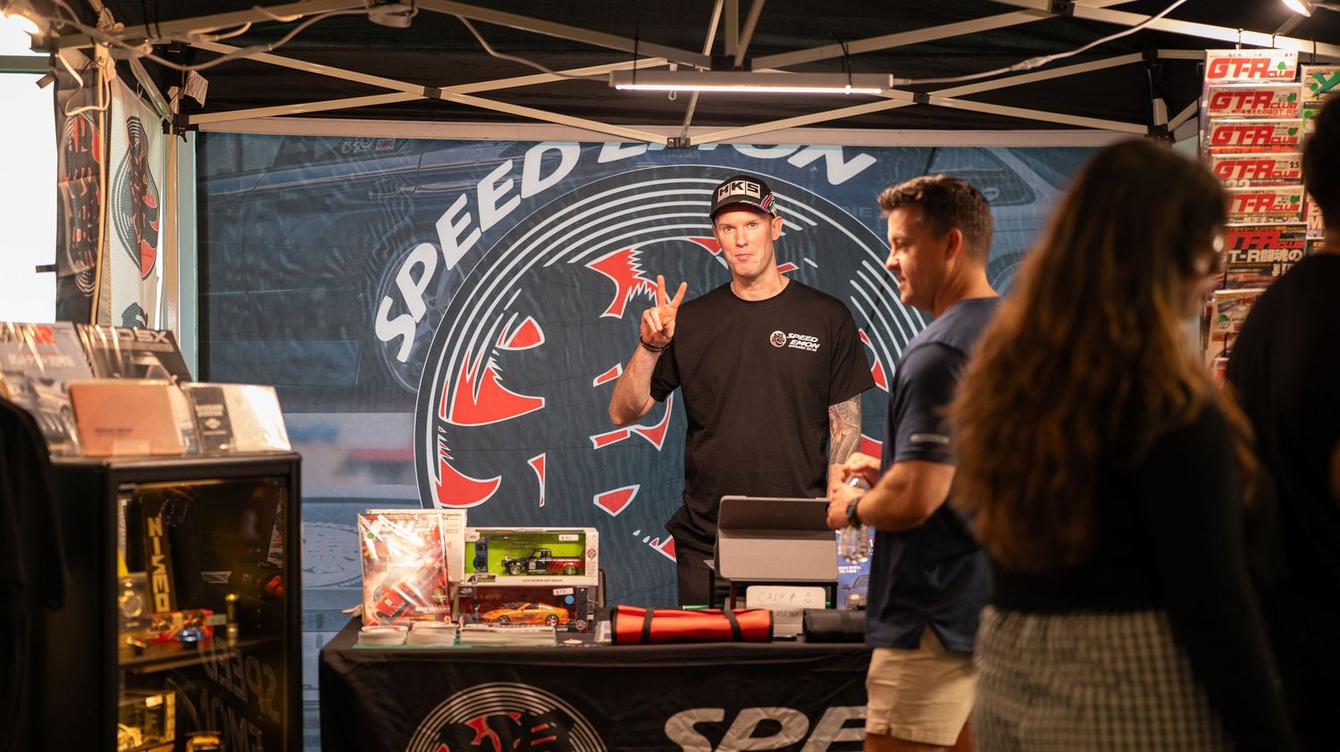 Car enthusiasts gather at a local speed shop with vintage and modern vehicles on display.