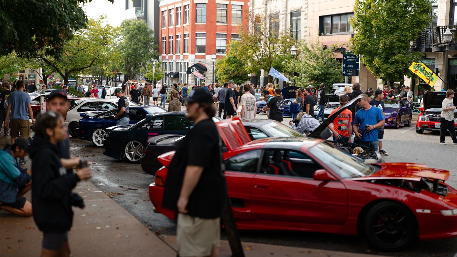 Red brick buildings line a street filled with parked cars and spectators at an auto show.