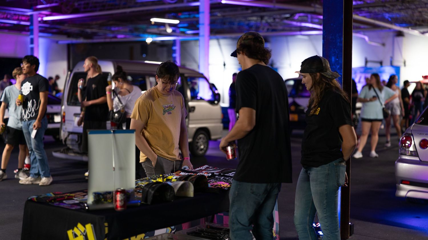 Indoor car meet with purple ambient lighting showing modified vehicles and attendees.
