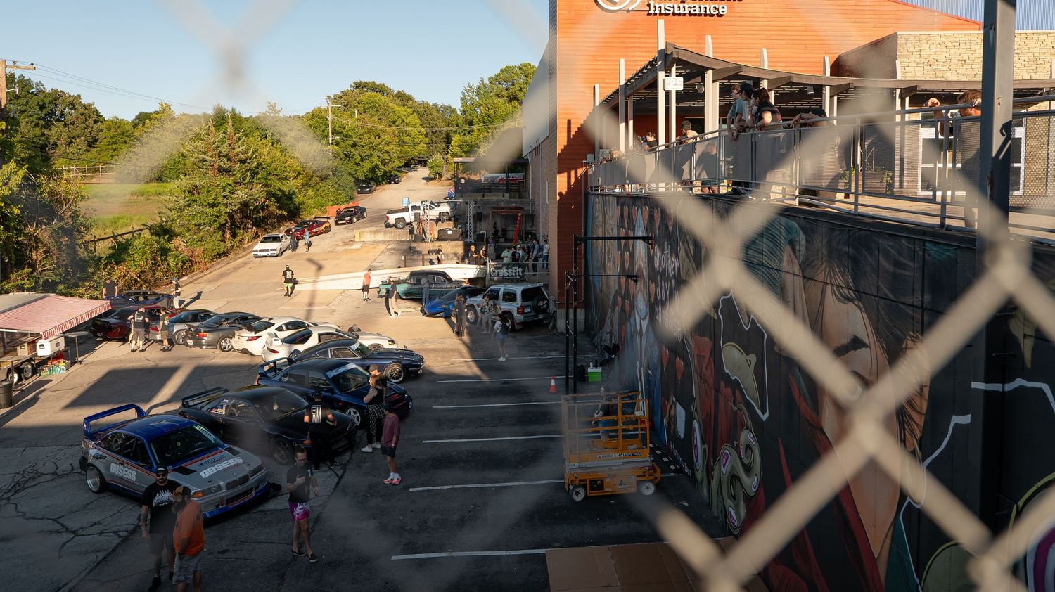 Panoramic view of a car meet through chain-link fence with orange building visible.