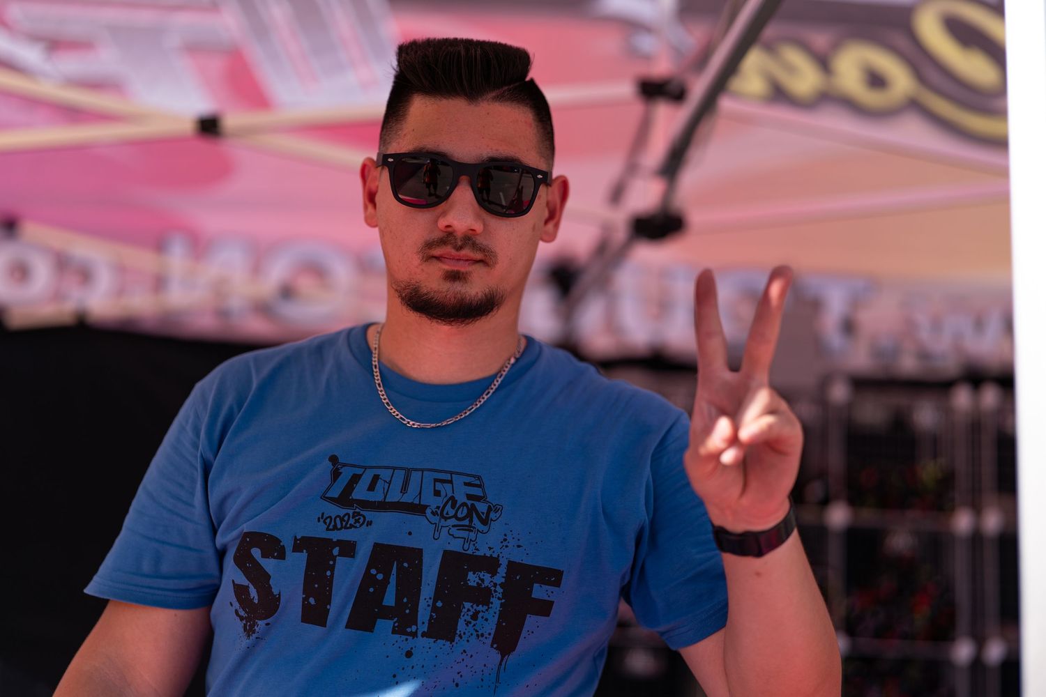Event staff member in blue t-shirt making peace sign while wearing sunglasses at outdoor venue.