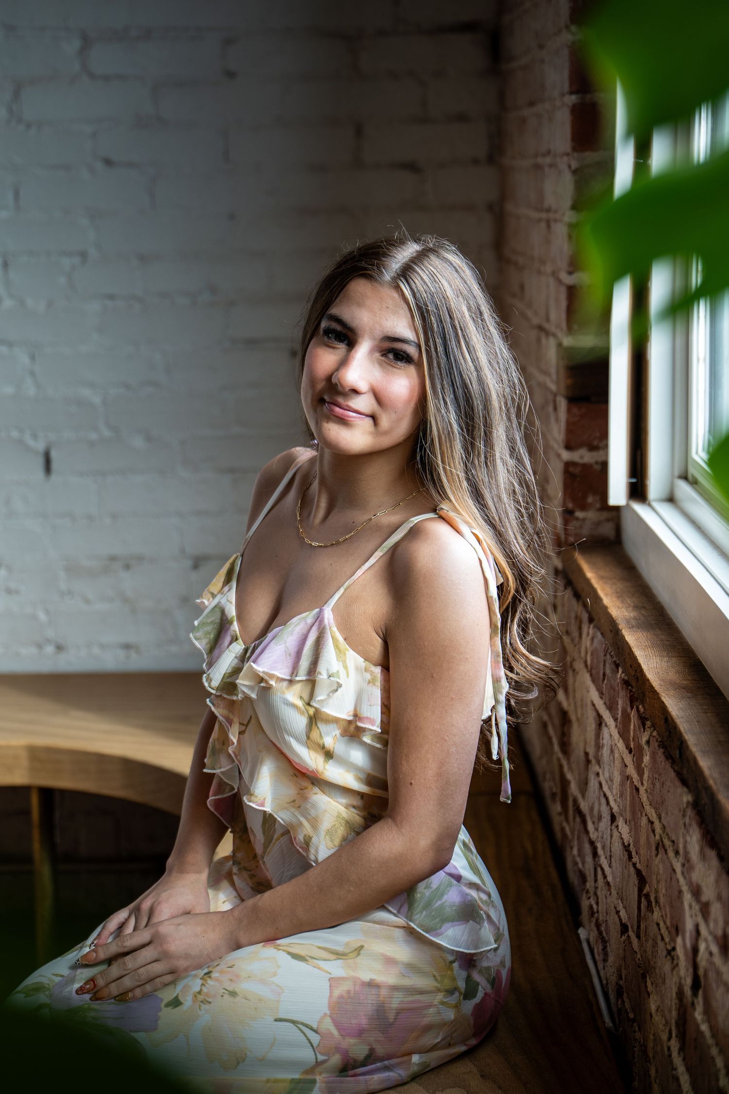 Young woman in floral ruffle dress sits smiling by brick wall window with green plant.