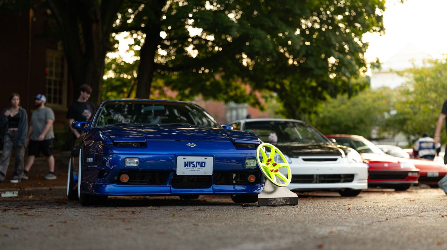 Street scene showing multiple modified performance cars parked under leafy trees.