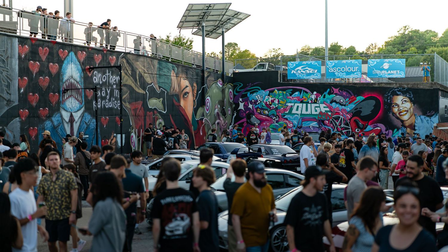 Panoramic view of a car show with colorful murals and gathered crowds.