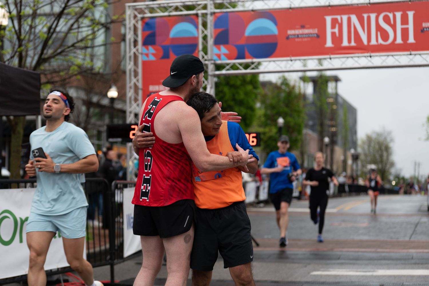 Runners embrace near the finish line of a race on a city street, celebrating their achievement on a cloudy day.