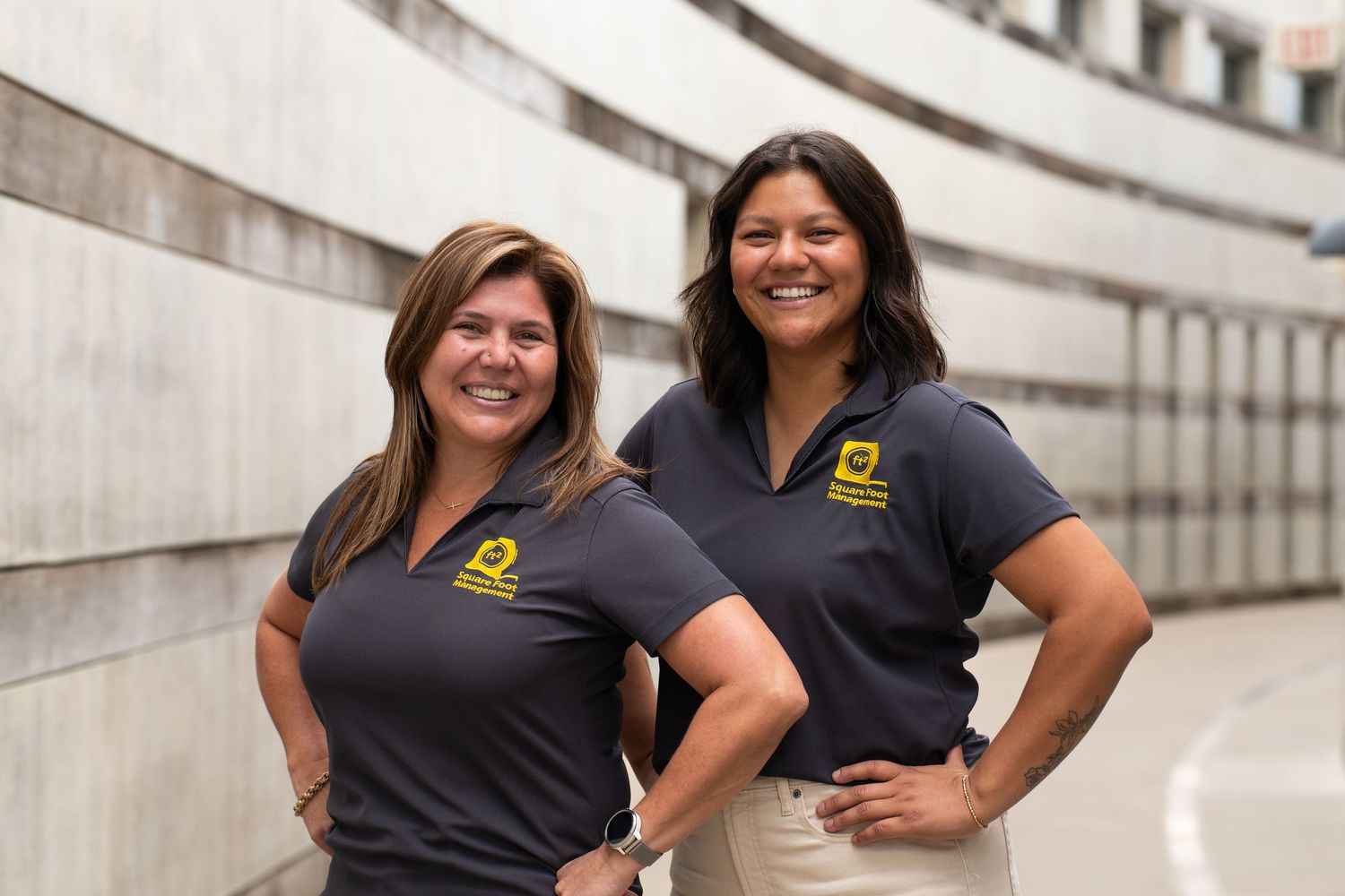 Two women in matching dark polo shirts with yellow logos smile confidently in a modern building hallway.