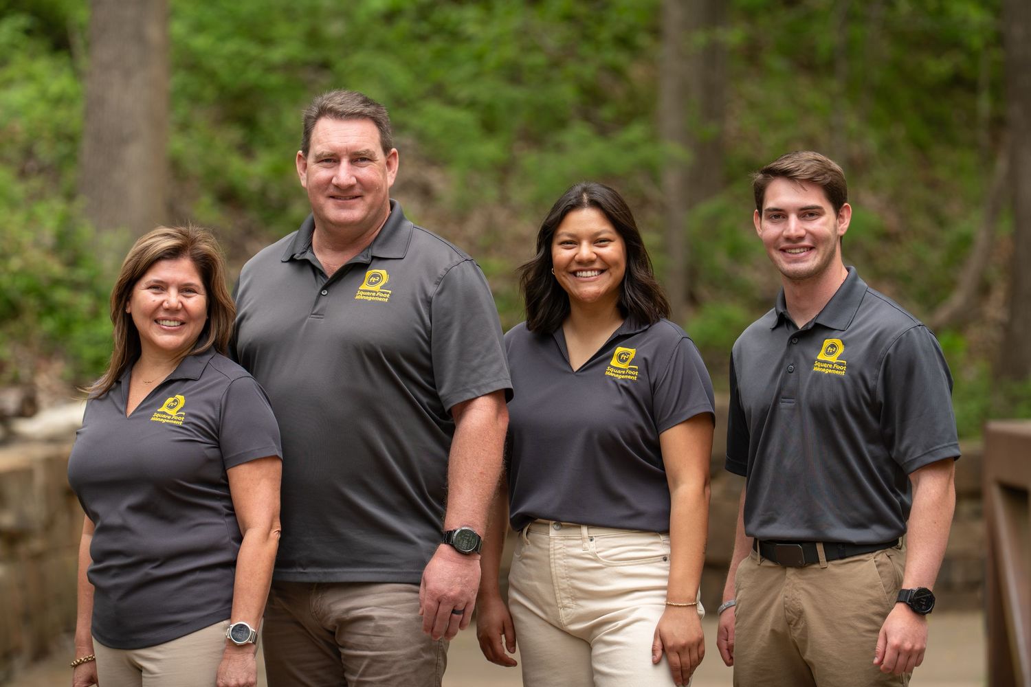 Four professionals in matching gray and yellow logo polo shirts smiling outdoors in a wooded setting.