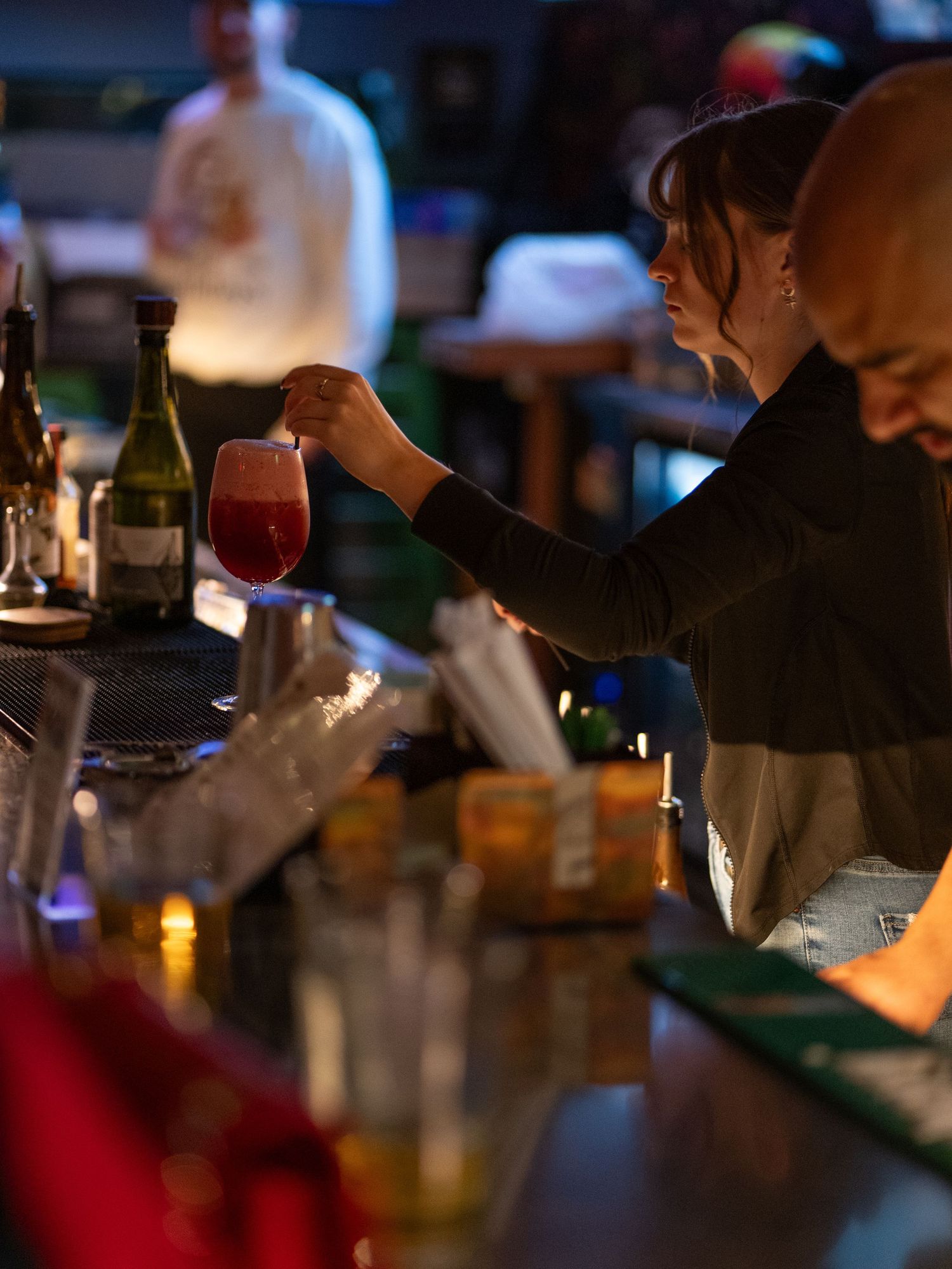 Dimly lit bar scene showing multiple patrons seated at counter with drinks and ambient lighting creating moody atmosphere.