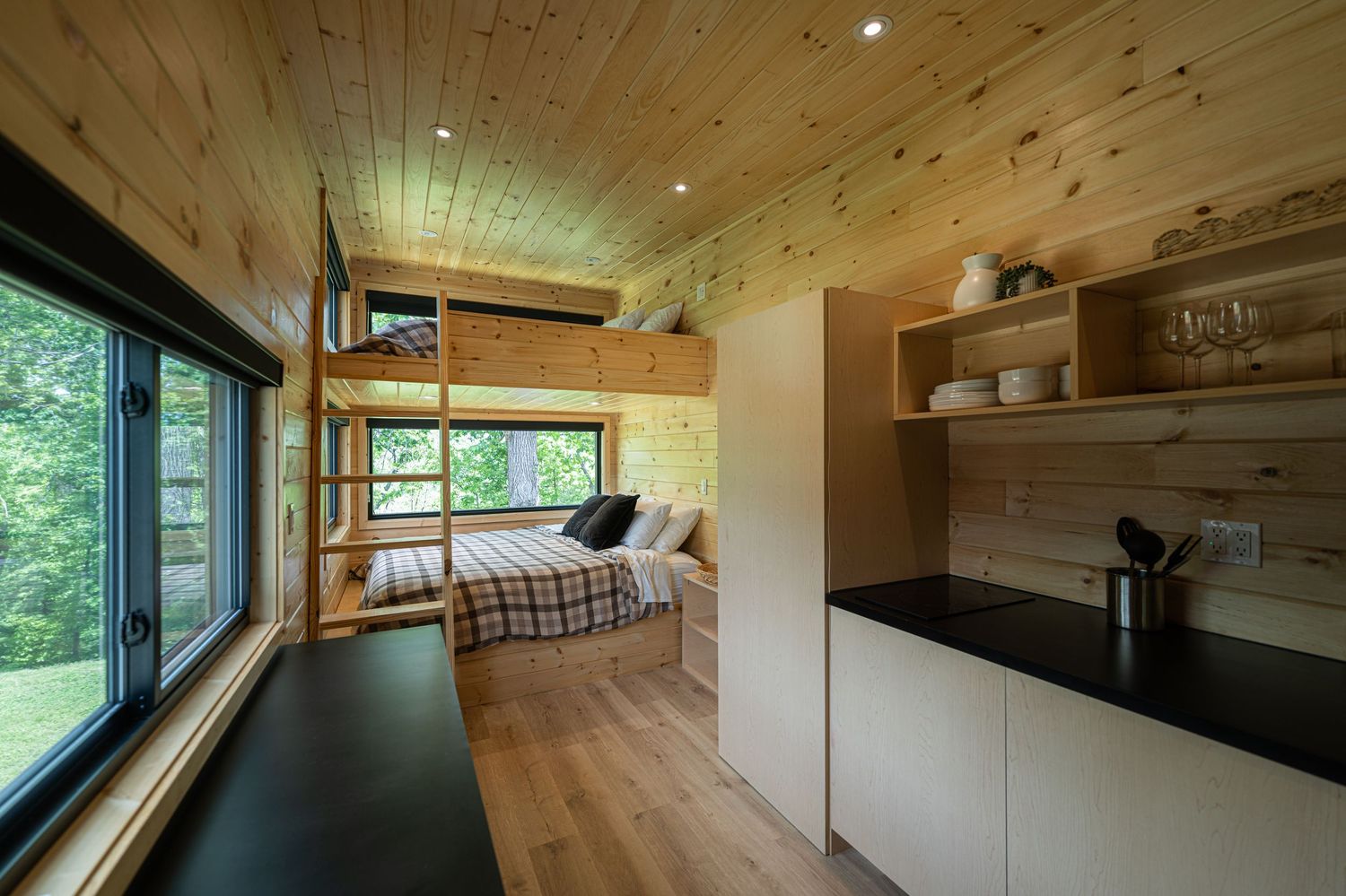 Cozy tiny home interior with bunk beds, wood-paneled walls, large windows, and a modern kitchenette with black countertops.
