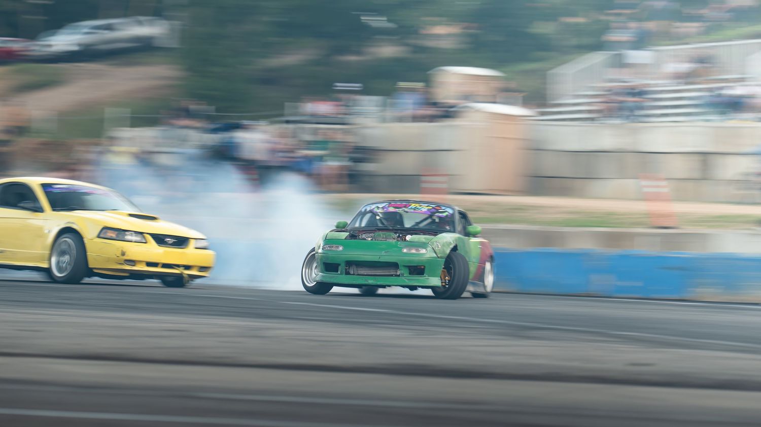 Multiple drift cars performing synchronized maneuvers on an outdoor track with tire smoke.