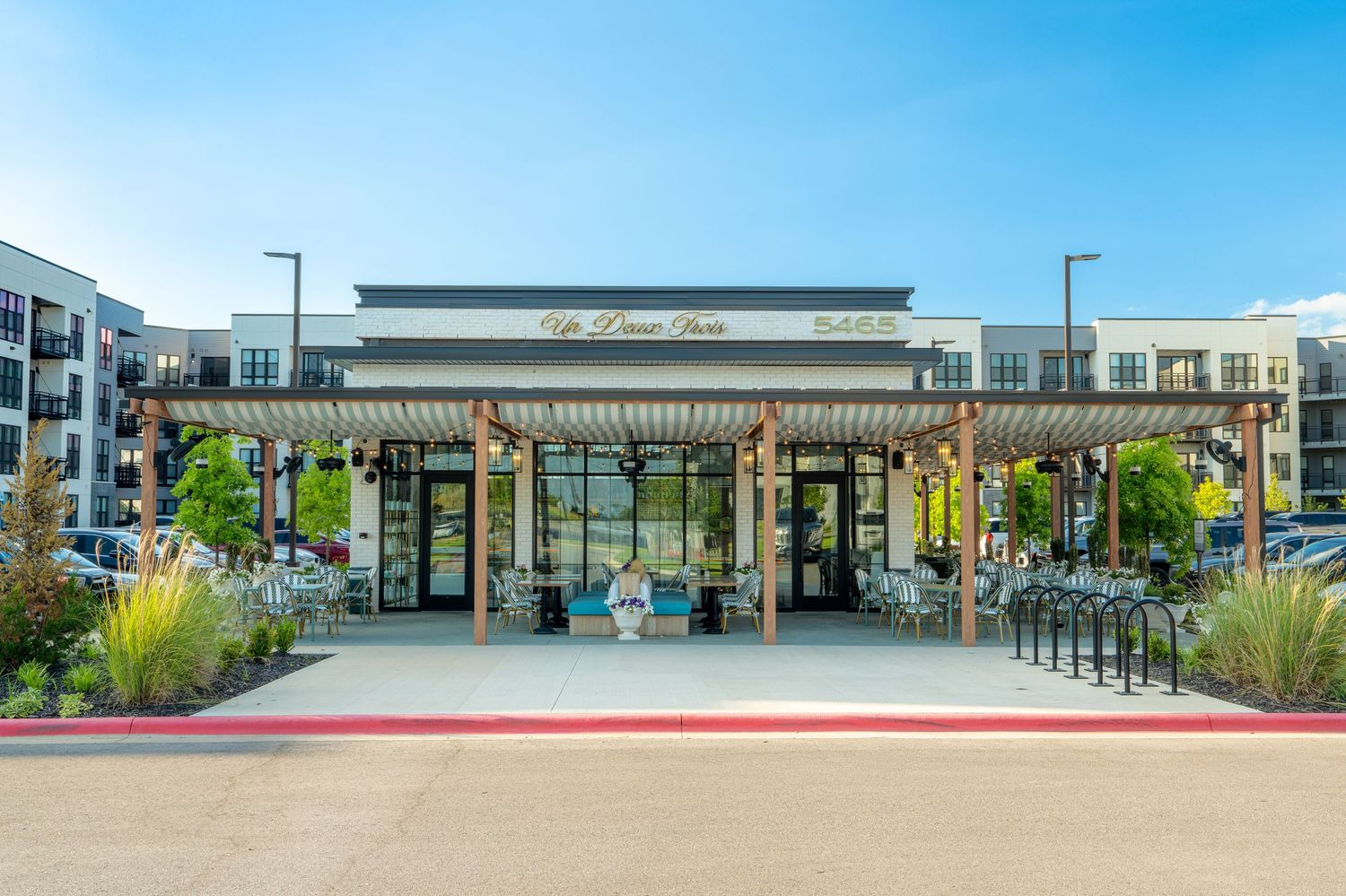 Modern retail storefront with pergola entrance, outdoor seating, and adjacent apartment complex under clear blue sky.