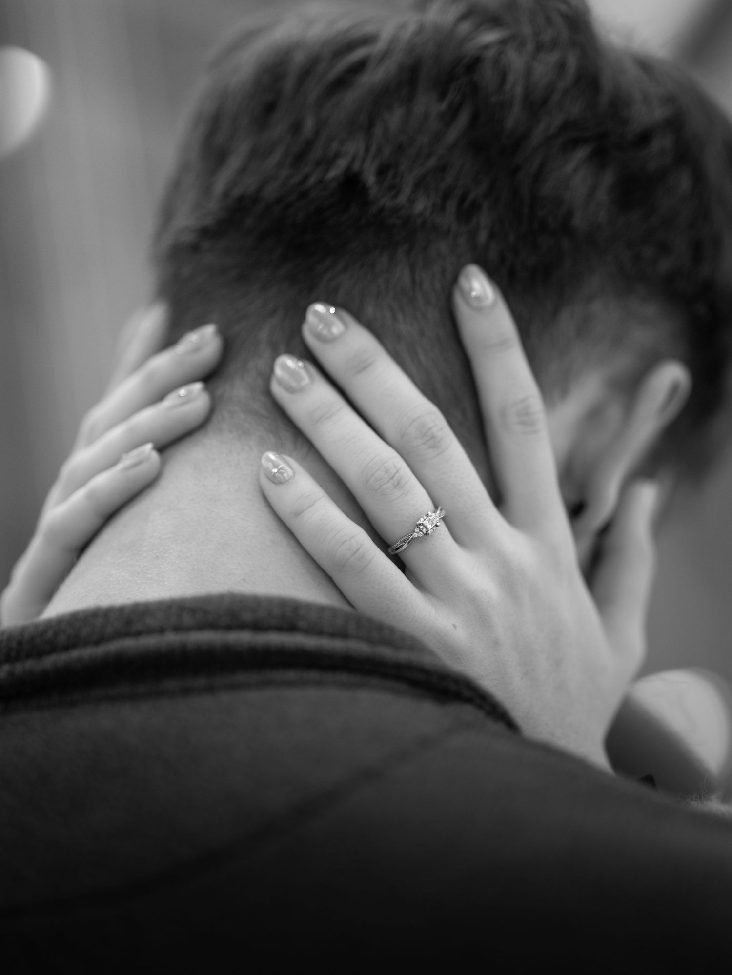 A close-up black and white photo shows hands with painted nails and an engagement ring embracing a neck from behind.