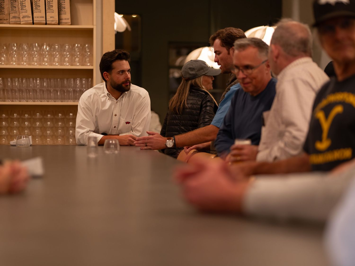 People gathered around a bar counter with wine glasses displayed on shelves in the background during an evening event.