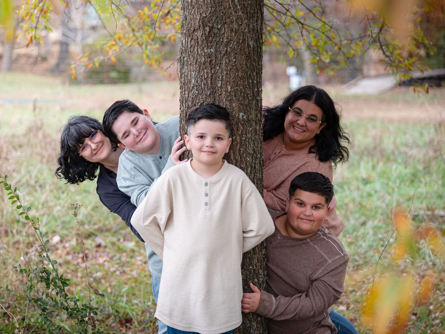 A playful group peeks out from behind a tree trunk in an autumn park setting with fallen leaves.