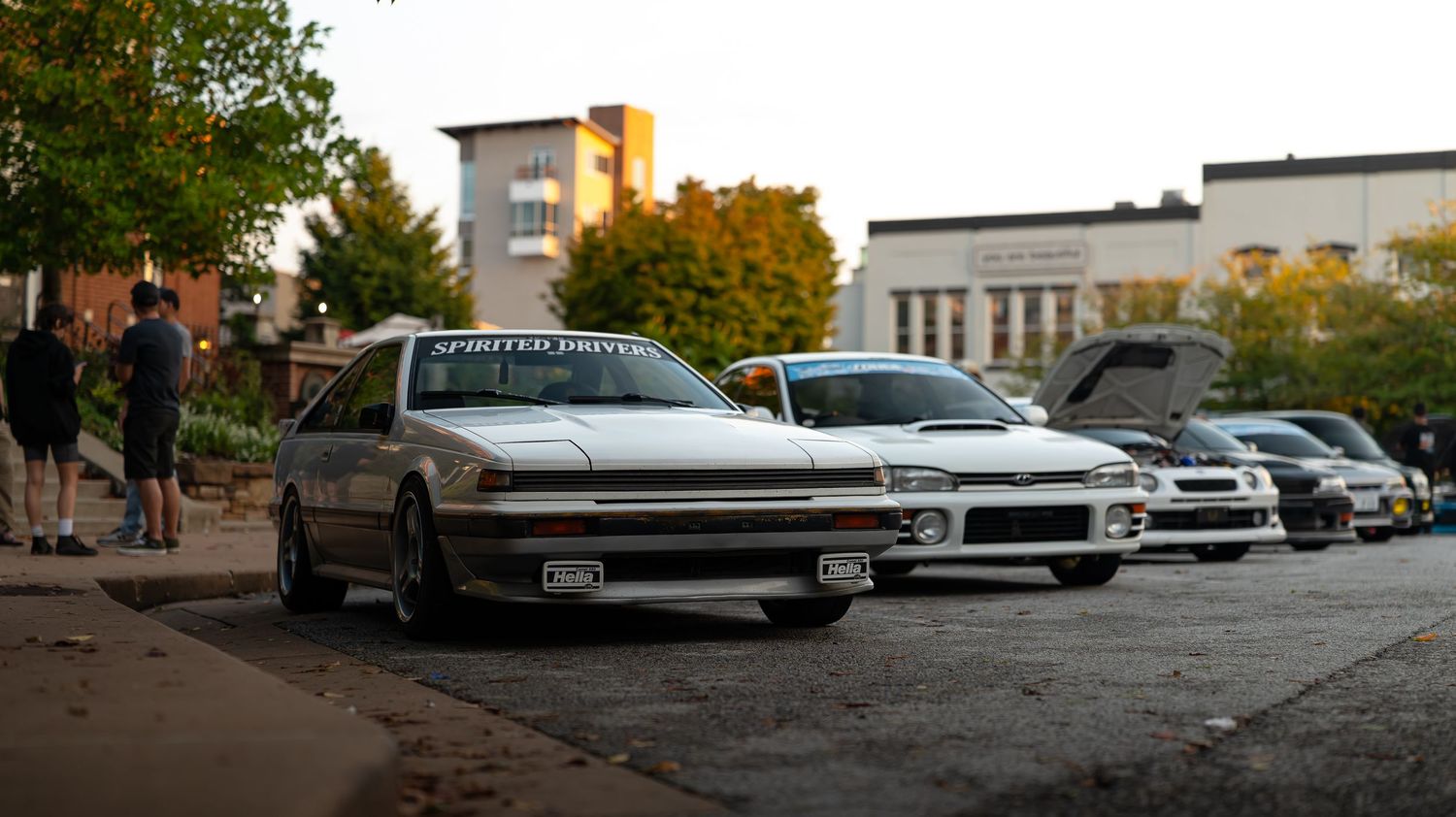 A row of modified sports cars parked at a car meet during sunset with trees in the background.