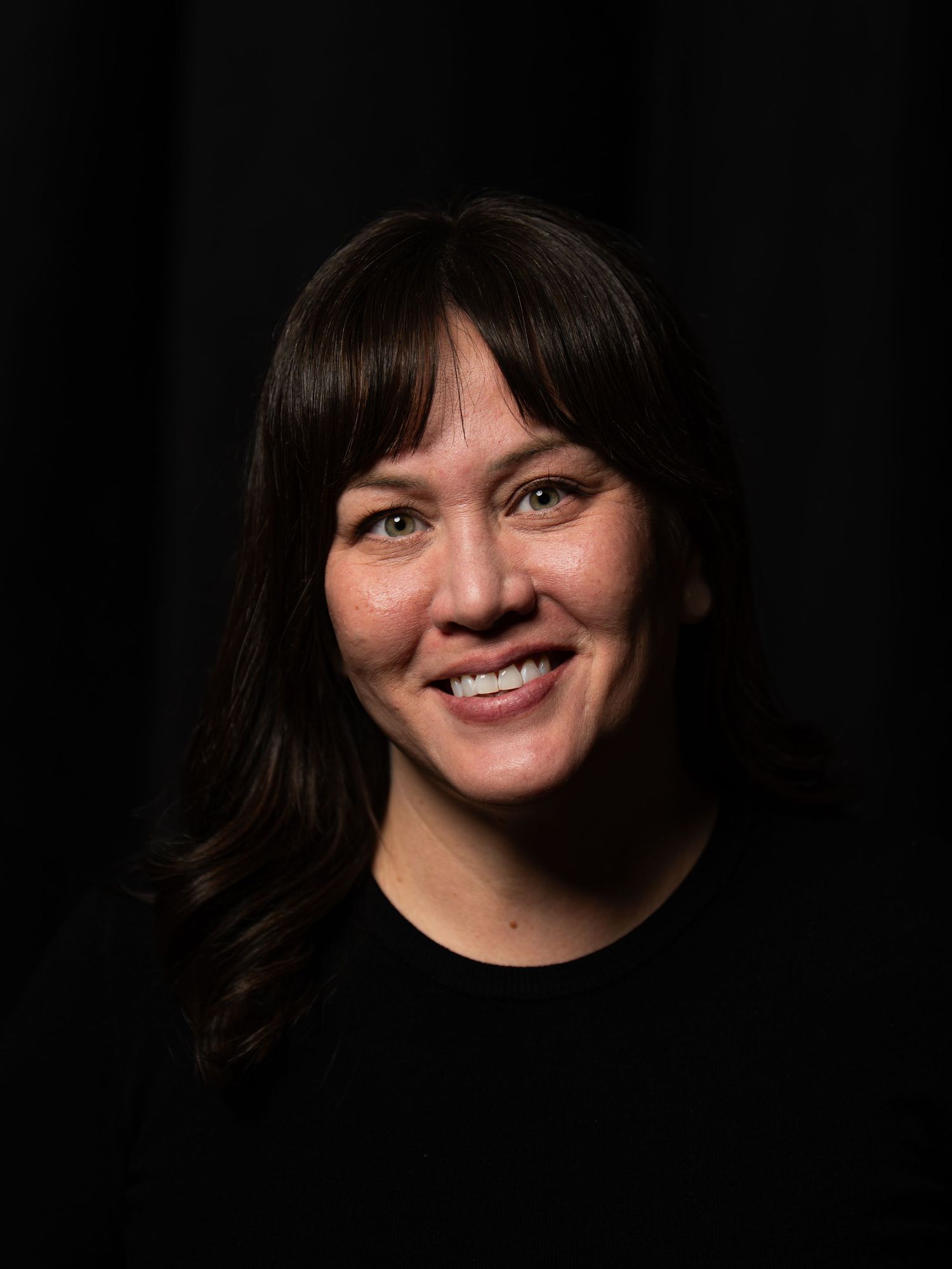 Professional headshot portrait of a person with dark hair and a warm smile against a black background.