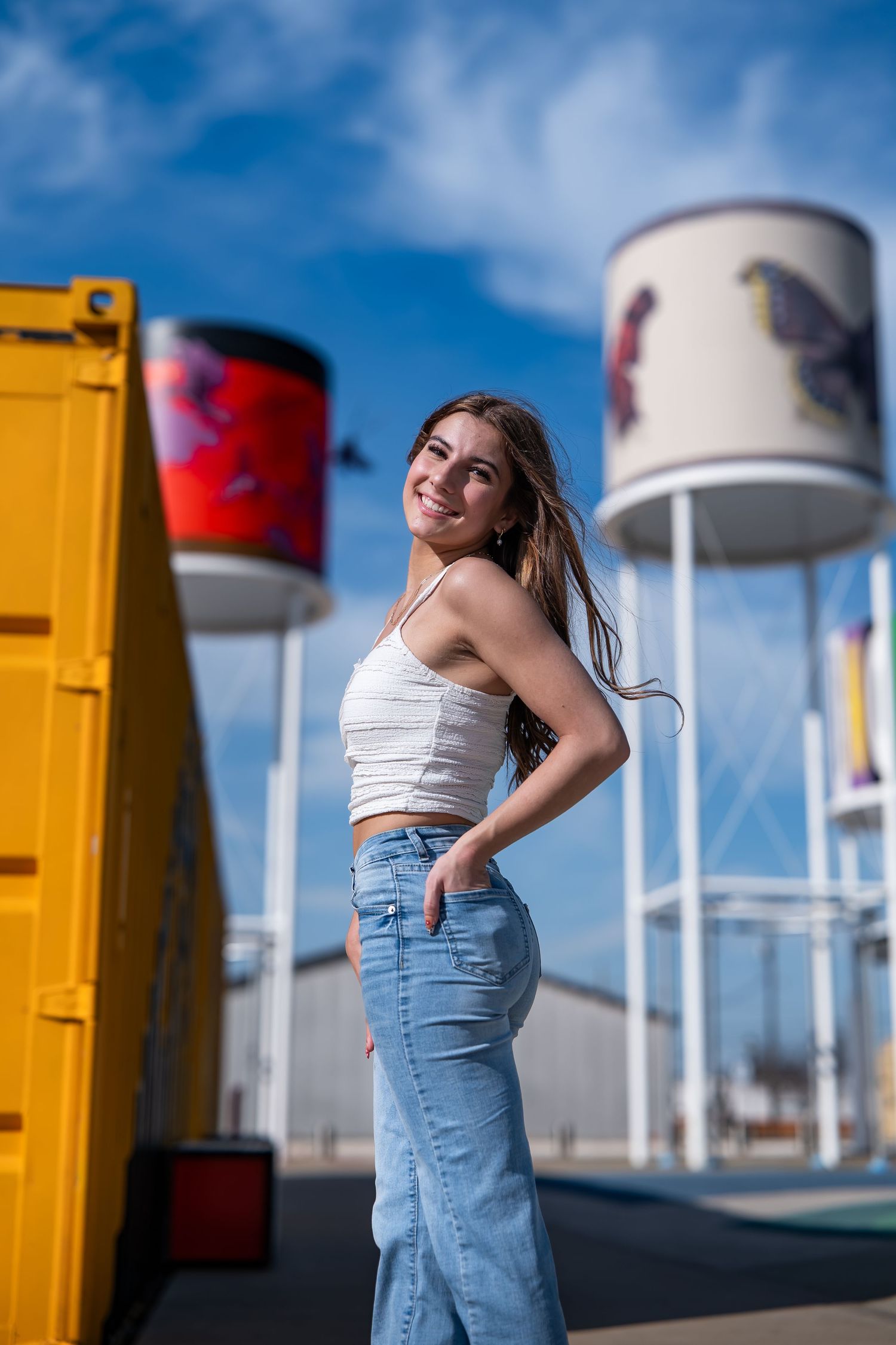 Young woman in white crop top and jeans smiling outdoors near colorful water towers under blue sky.