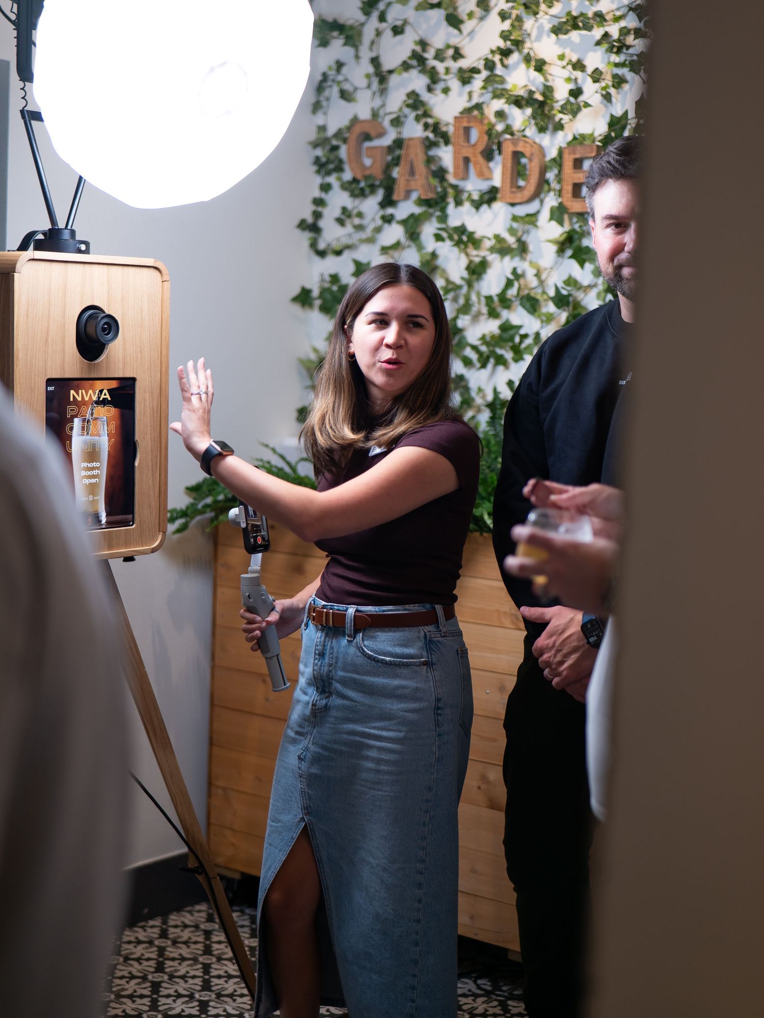 Someone in a black t-shirt and denim skirt poses against a white wall with greenery decor in a sequence of photos.