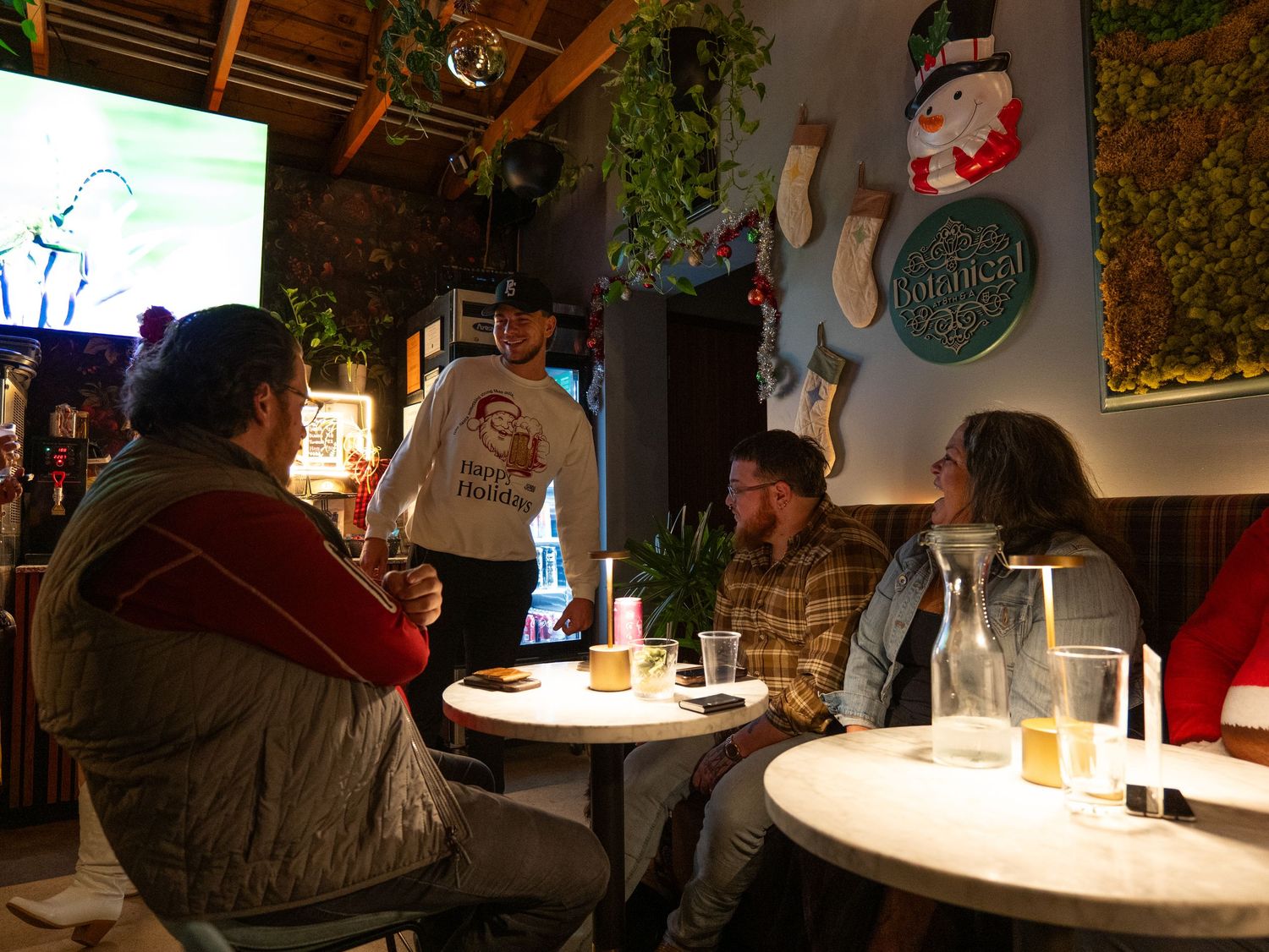 Patrons sit at illuminated tables inside a dimly lit bar with exposed brick walls and hanging plants.