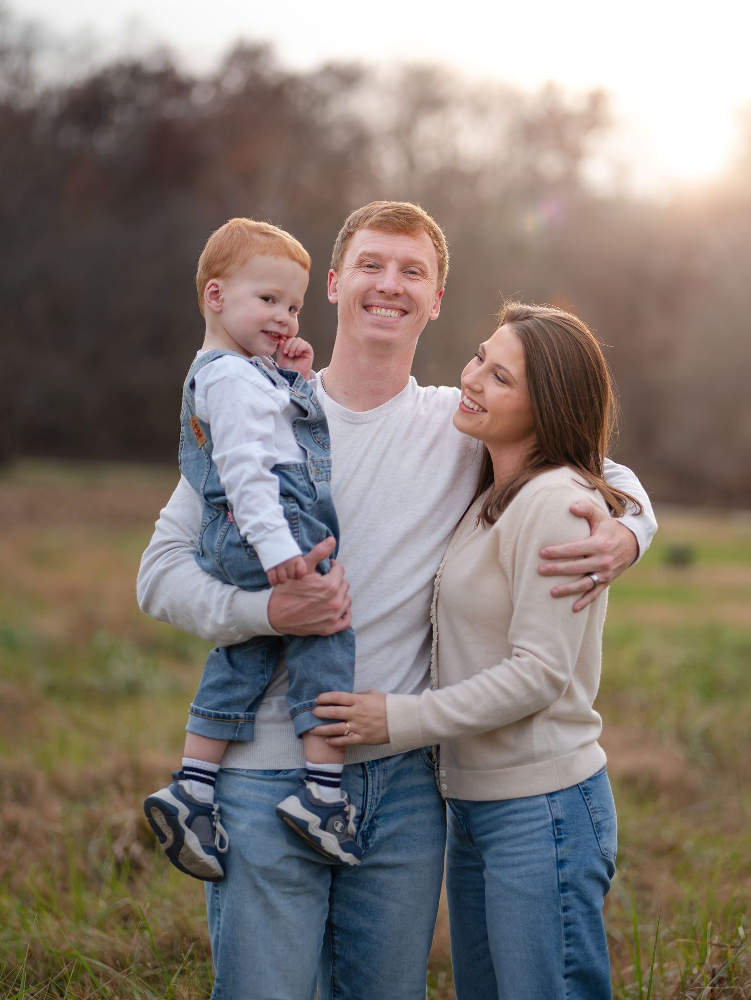 A family shares tender moments together in a field during golden hour as sunlight filters through the trees.