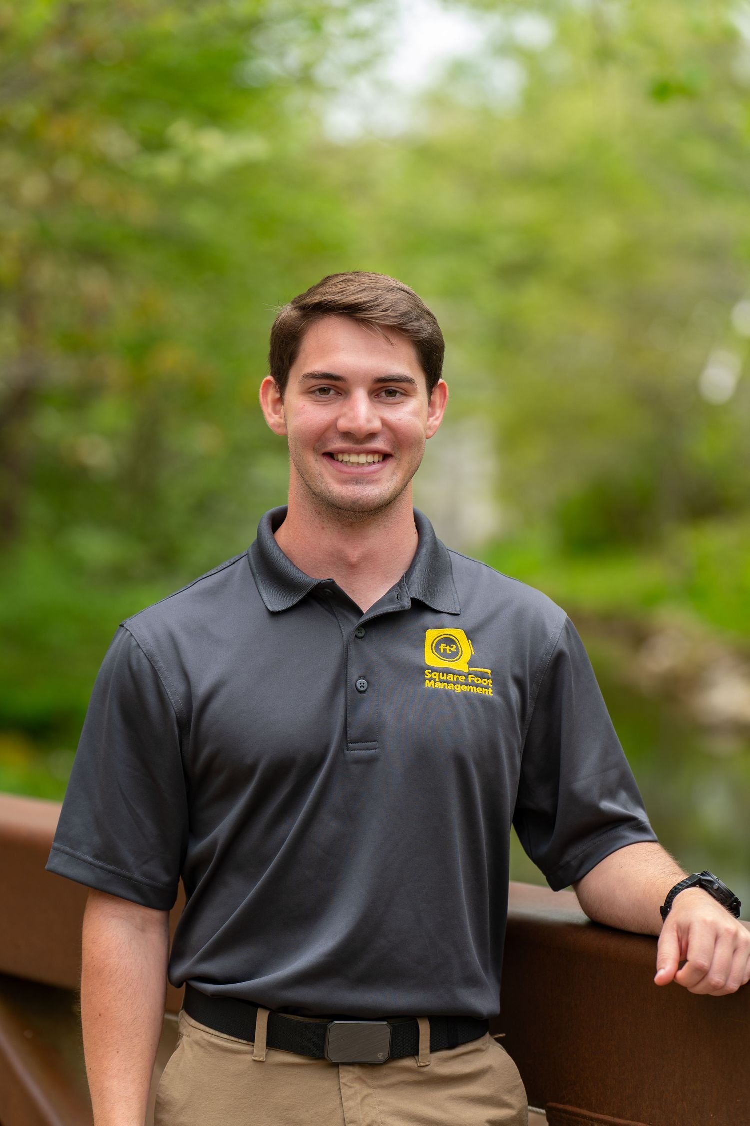 Young man in gray polo shirt smiling outdoors on a wooden bridge surrounded by lush green trees.