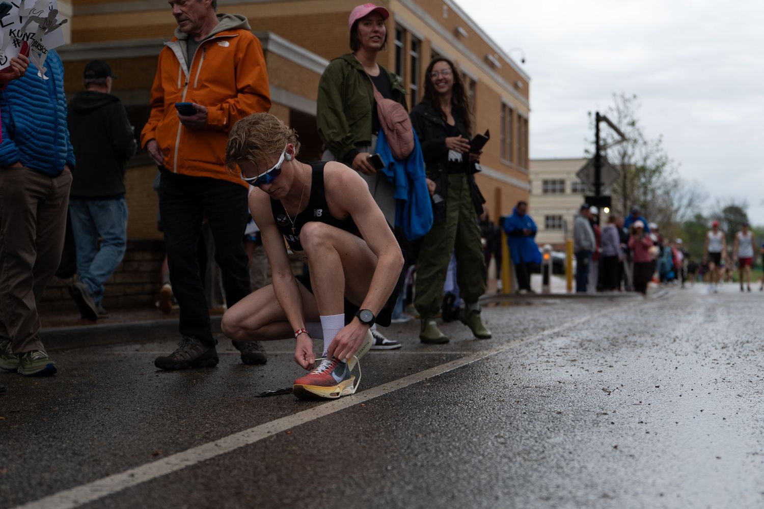 Runner tying shoelaces on wet pavement during a road race, surrounded by spectators on a rainy day.