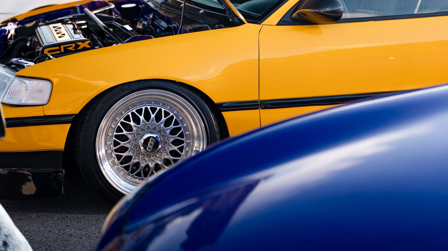 Close-up of yellow and blue cars showing custom wheels at car meet.