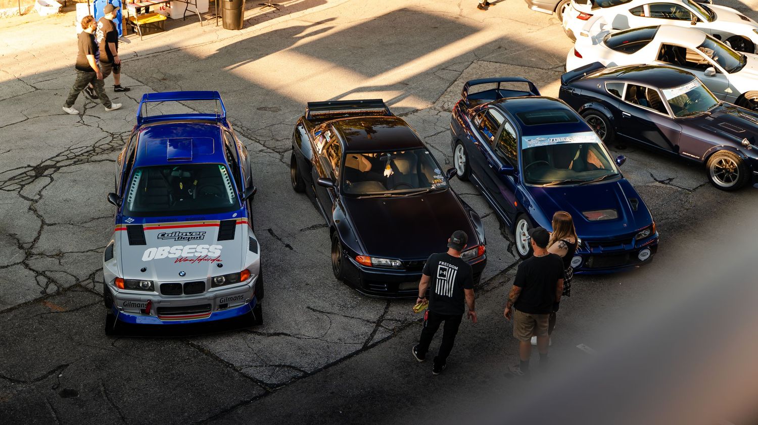 Cars parked in a row at a car meet with people gathering around.