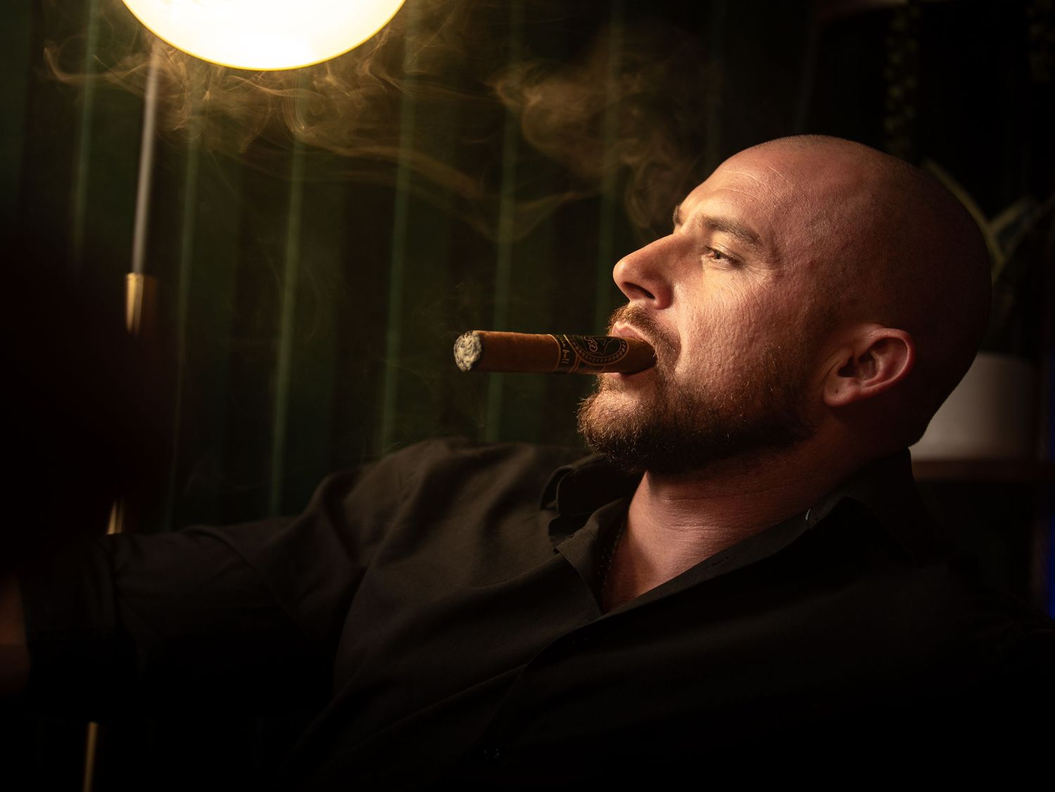 A person smoking a cigar in a dimly lit room with dramatic lighting from above casting shadows on their face.