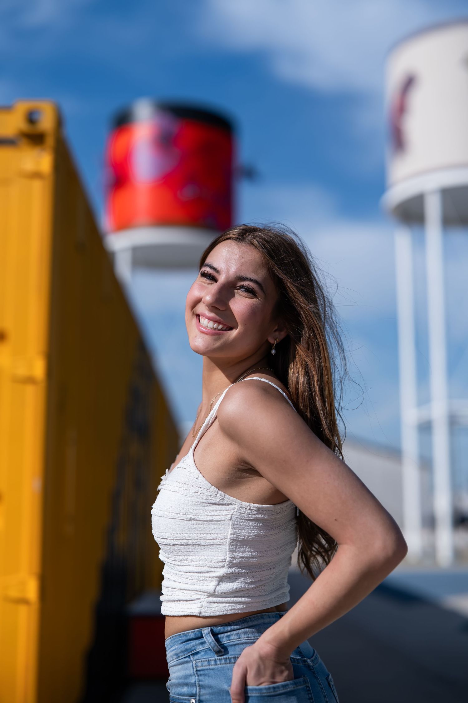 Smiling young woman in white crop top and jeans poses outdoors near colorful industrial structures under blue sky.