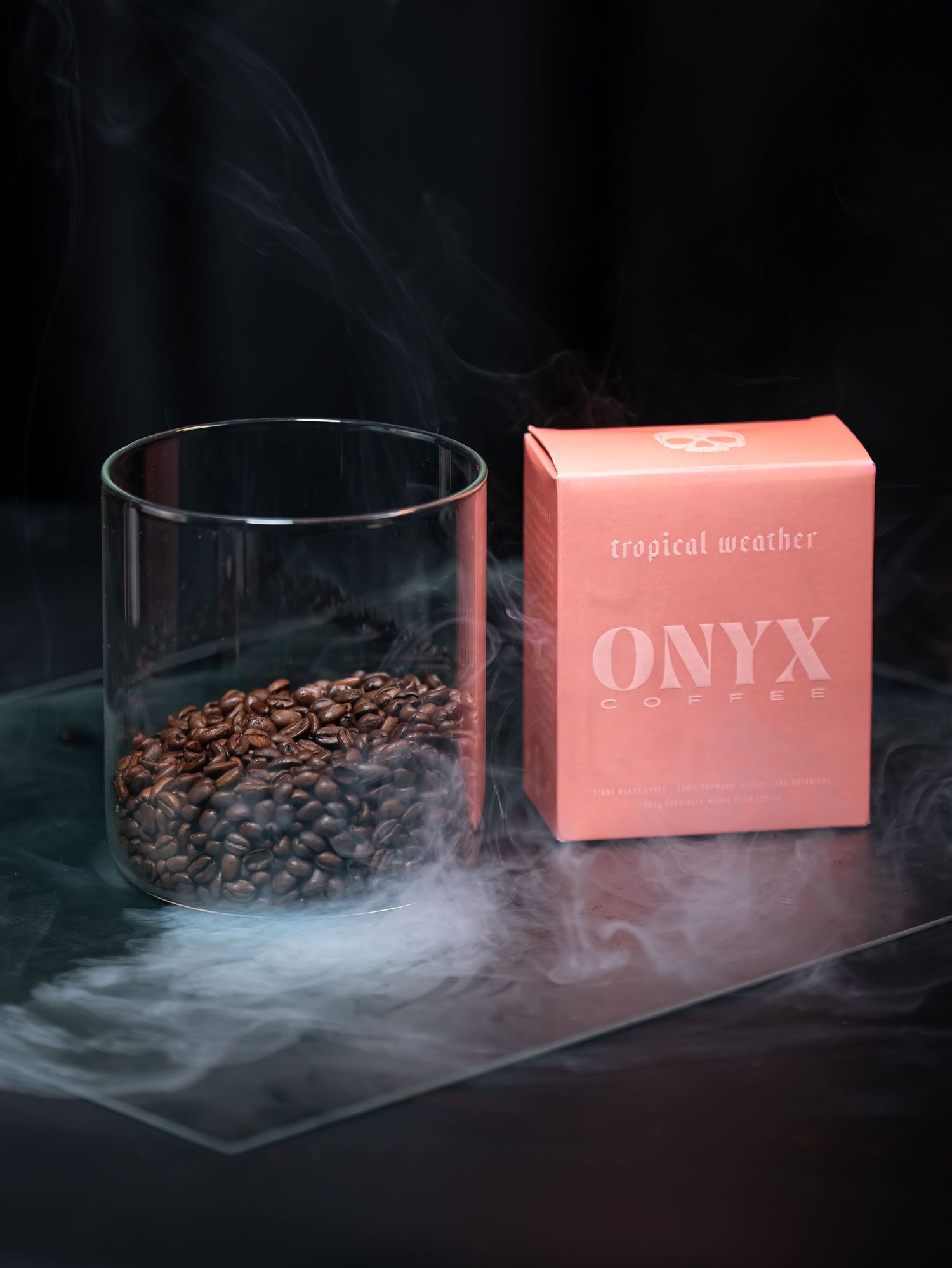 A glass container filled with coffee beans next to a pink Onyx product box with smoke effects in a dark setting.