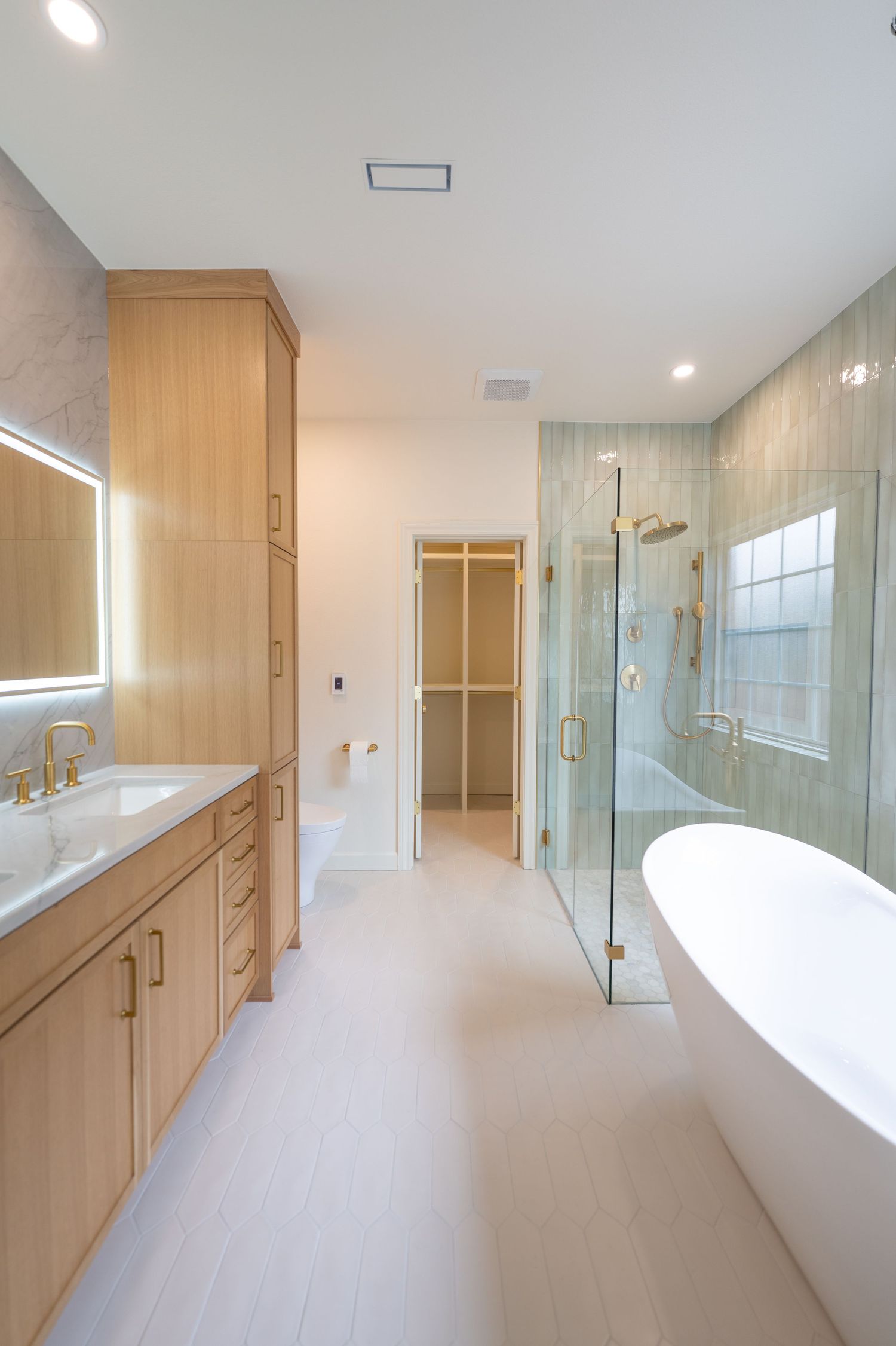 Modern luxury bathroom with wood vanity, freestanding soaking tub, glass shower, and gold fixtures on white hardwood floors.