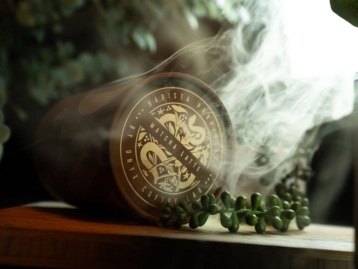 Aromatic wooden candle with smoke rising, adorned with decorative gold emblem, beside green botanicals on wooden surface.