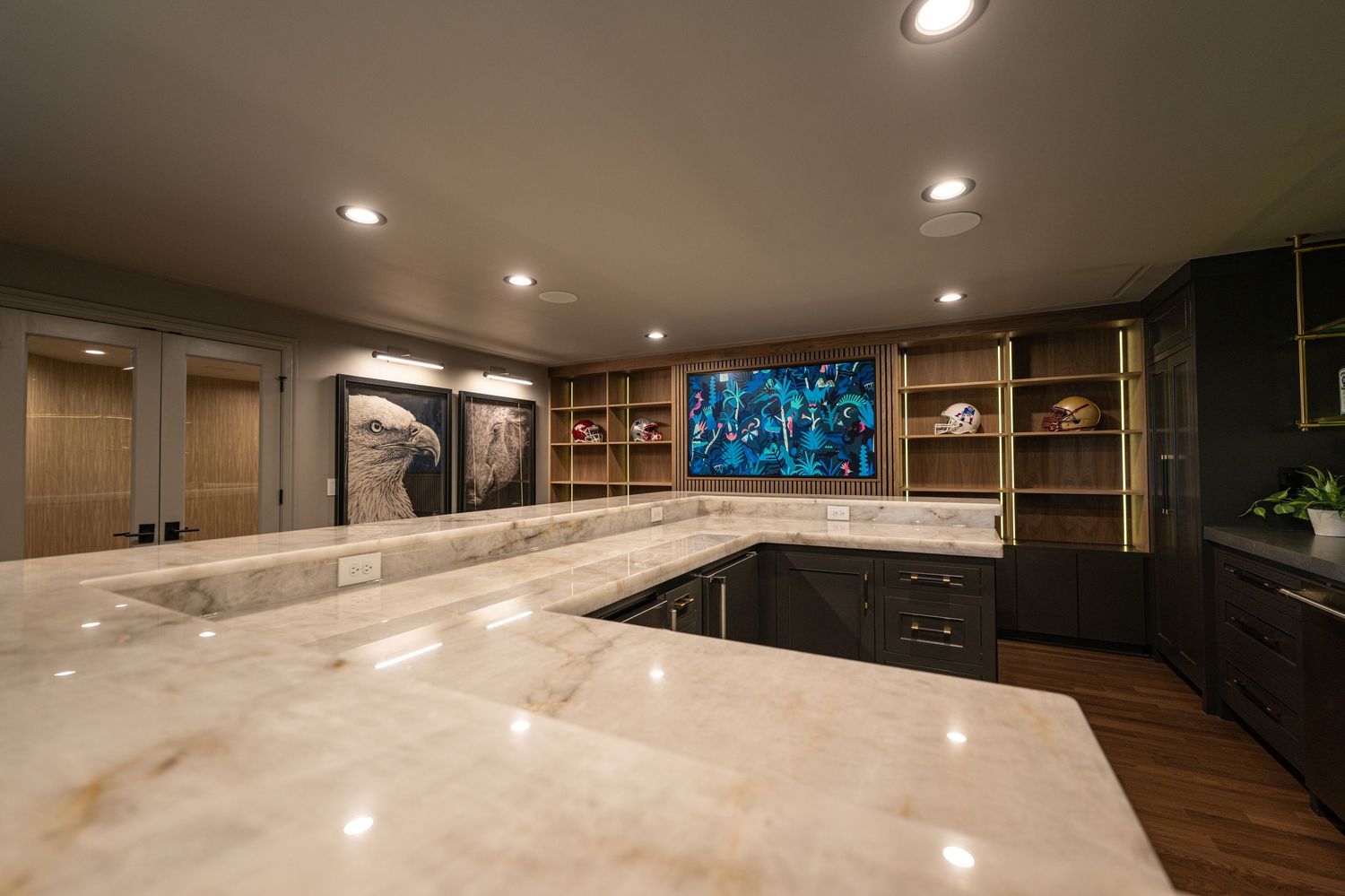 Luxury basement bar with marble countertops, built-in shelving, dark cabinetry, and a wall-mounted TV in an elegant entertainment space.