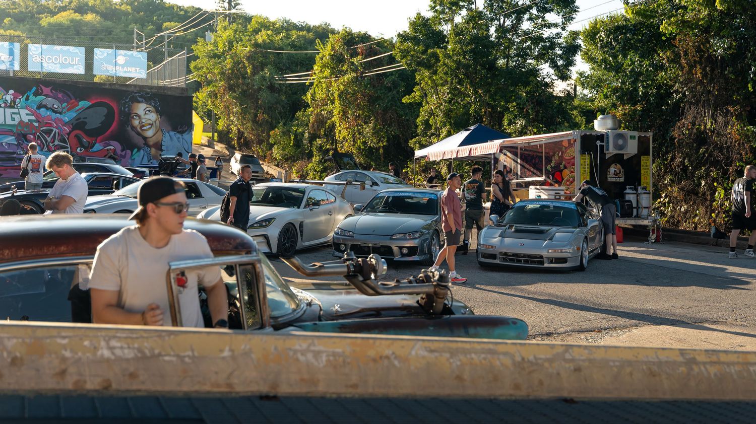 Car enthusiasts gather at a sunset meetup in a parking lot with modified vehicles on display.