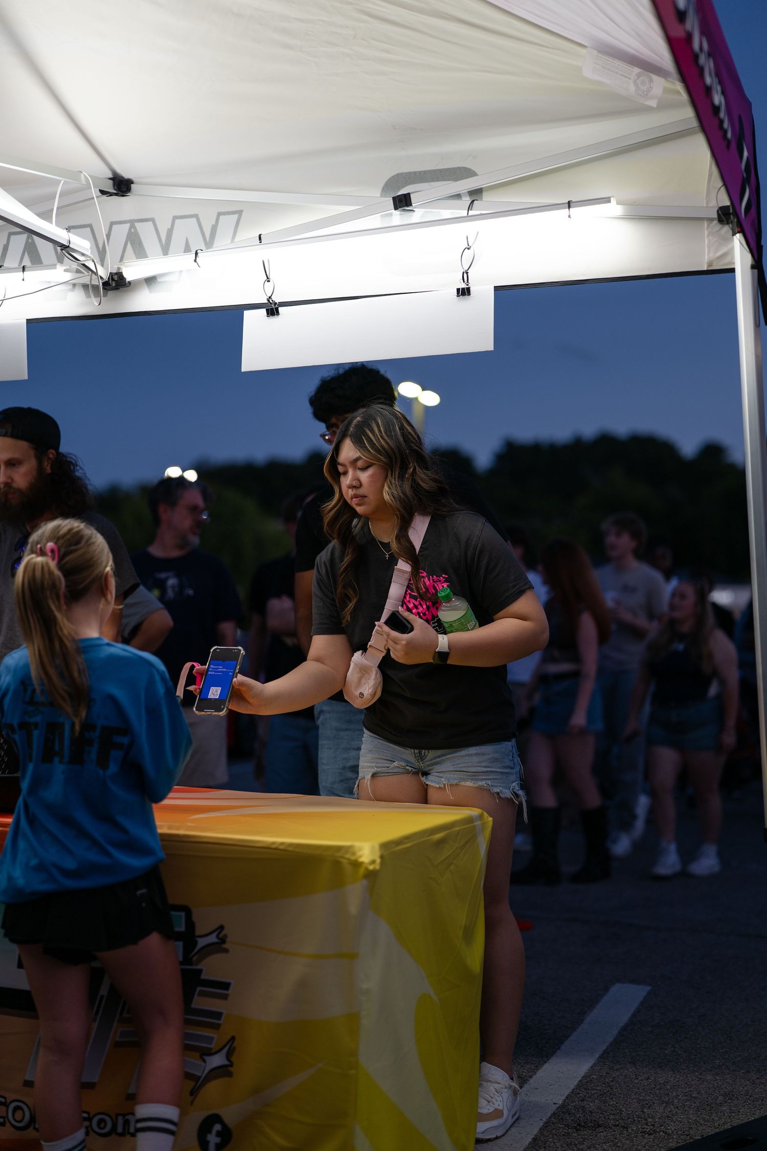 White tent setup with yellow counter and people gathering at dusk.