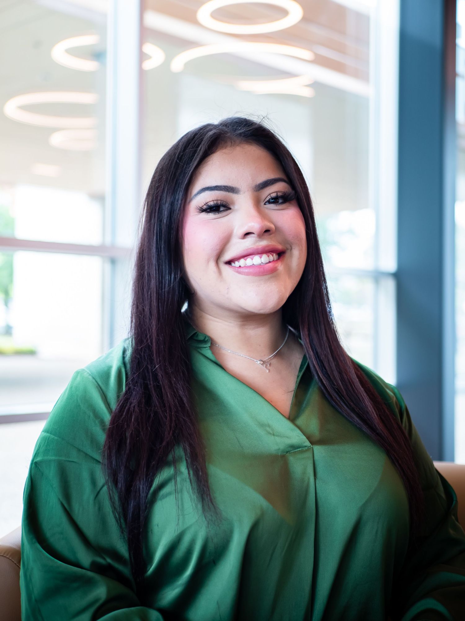 A person wearing a green blouse smiles while seated in an office setting with circular ceiling lights.