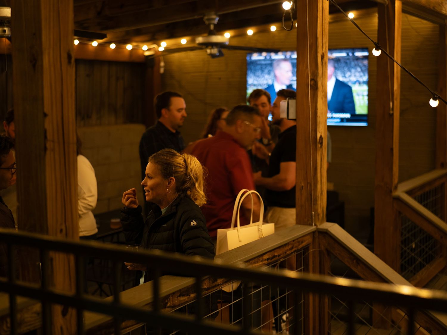 Customers gather inside a dimly lit restaurant bar with string lights and TV screens visible in the background.