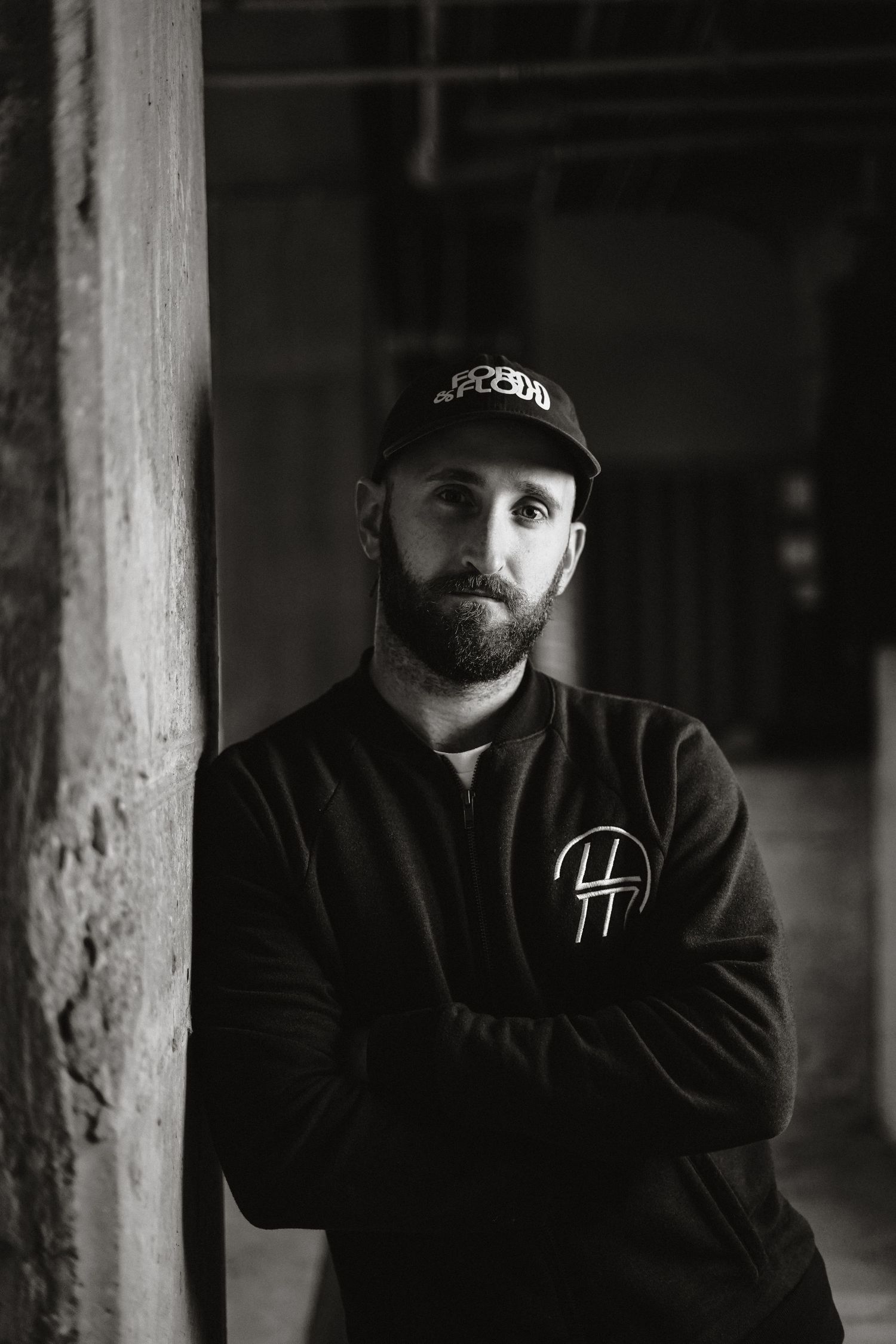 Black and white portrait of someone wearing a baseball cap and dark hoodie in a shadowy industrial setting.