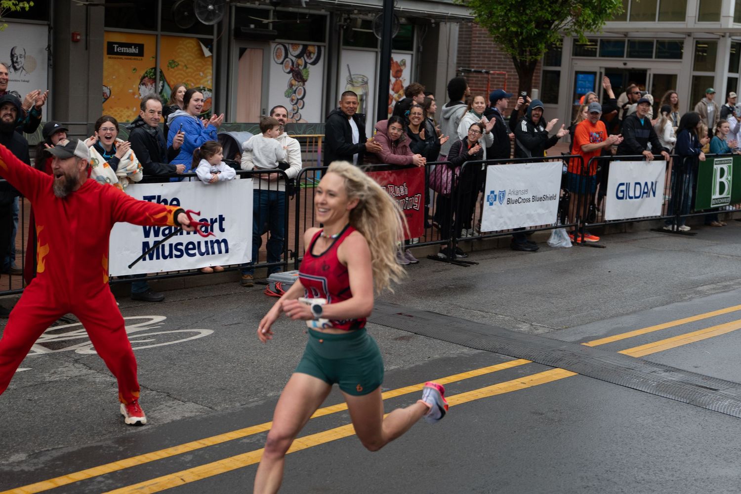Woman runner smiles crossing finish line at street race with cheering crowd and Gildan sponsor banner visible.