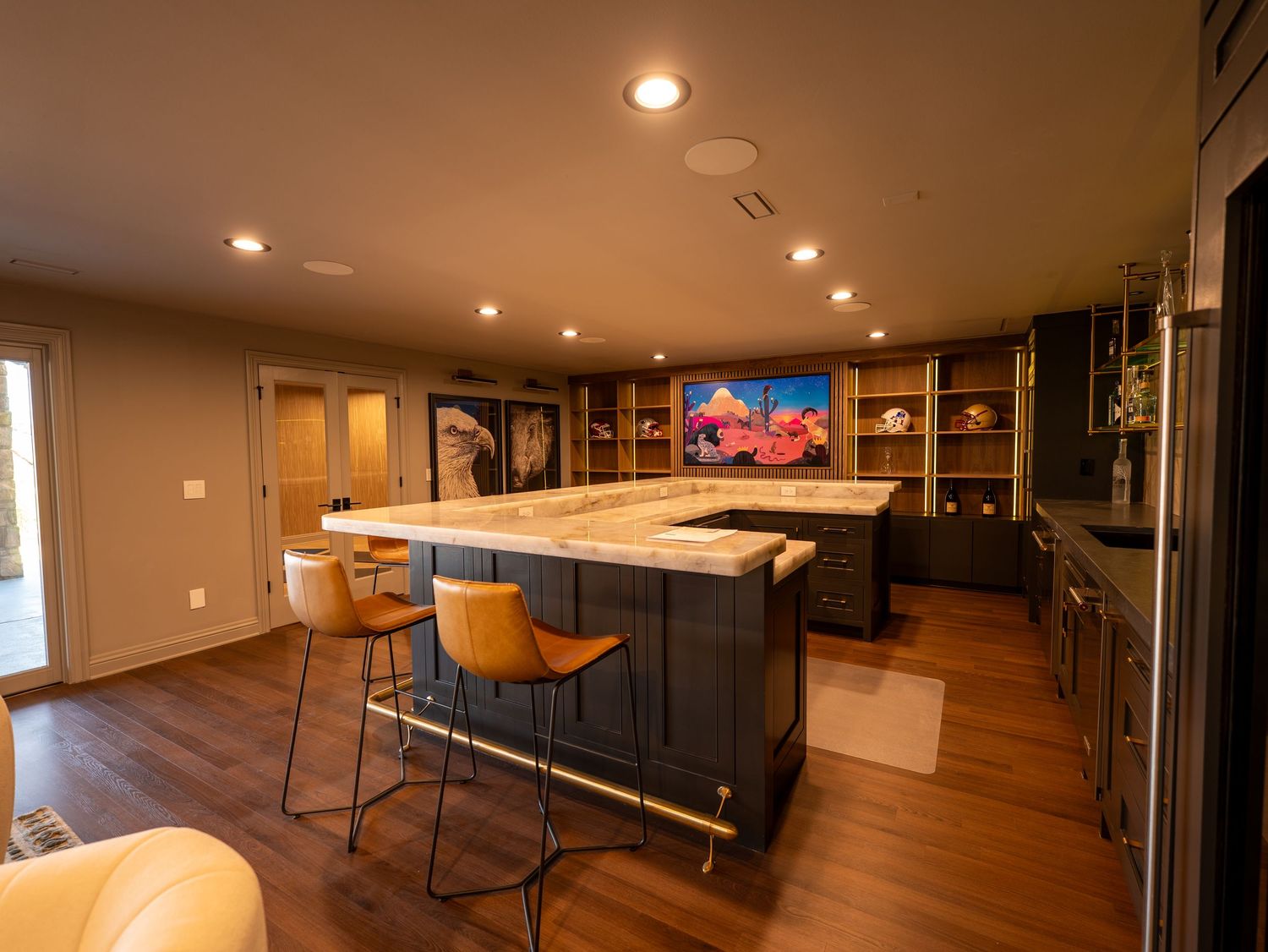 Spacious open-concept kitchen with large marble island, bar stools, dark cabinetry, hardwood floors, and warm recessed lighting.