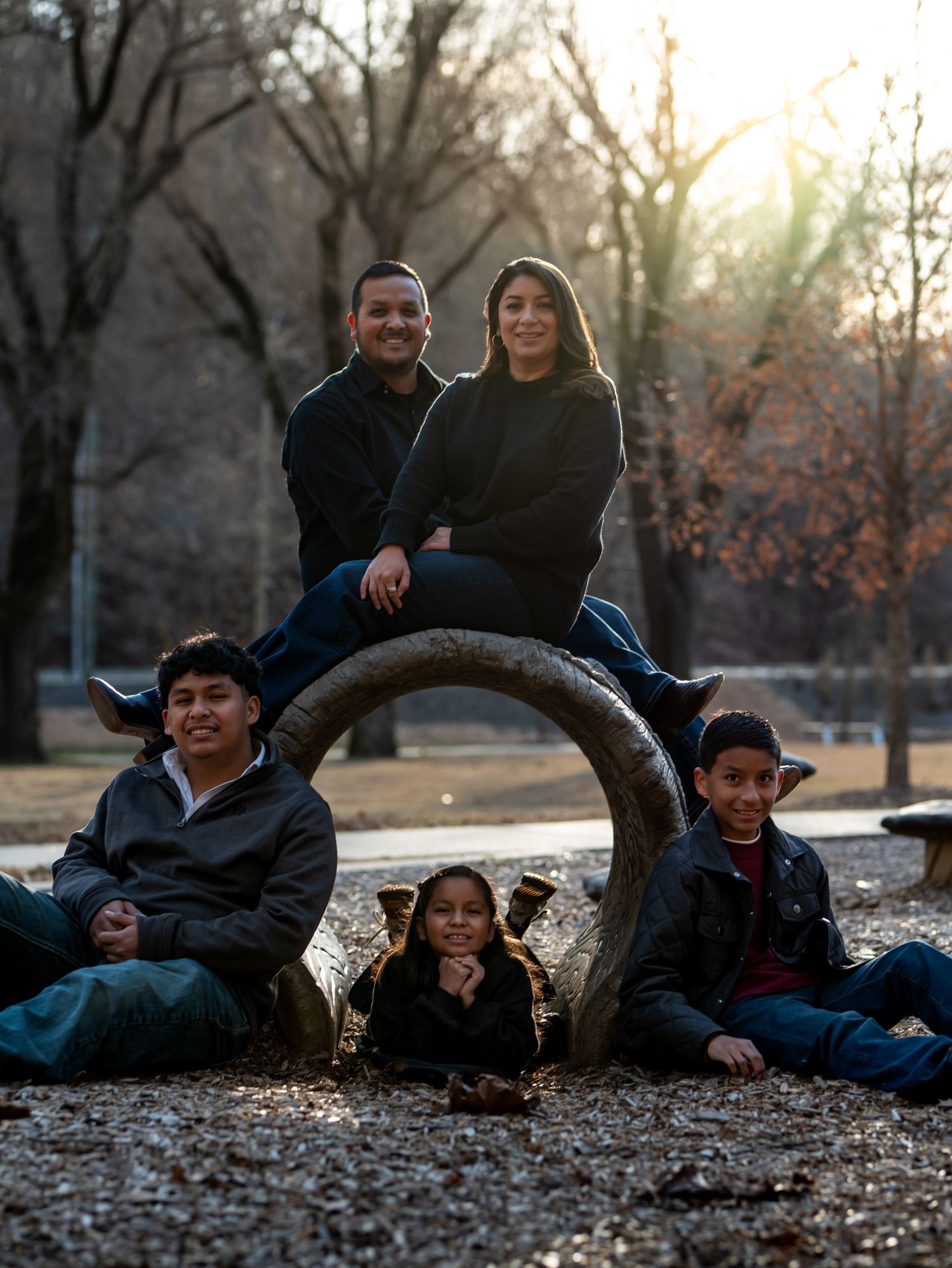 A family lounges together on fallen autumn leaves in a park during a casual outdoor photo session at sunset.