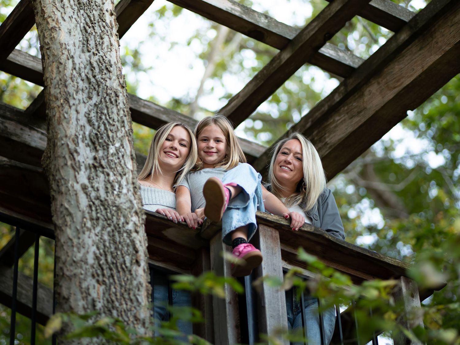 Three people lean over wooden beams of an outdoor structure surrounded by trees and natural lighting.