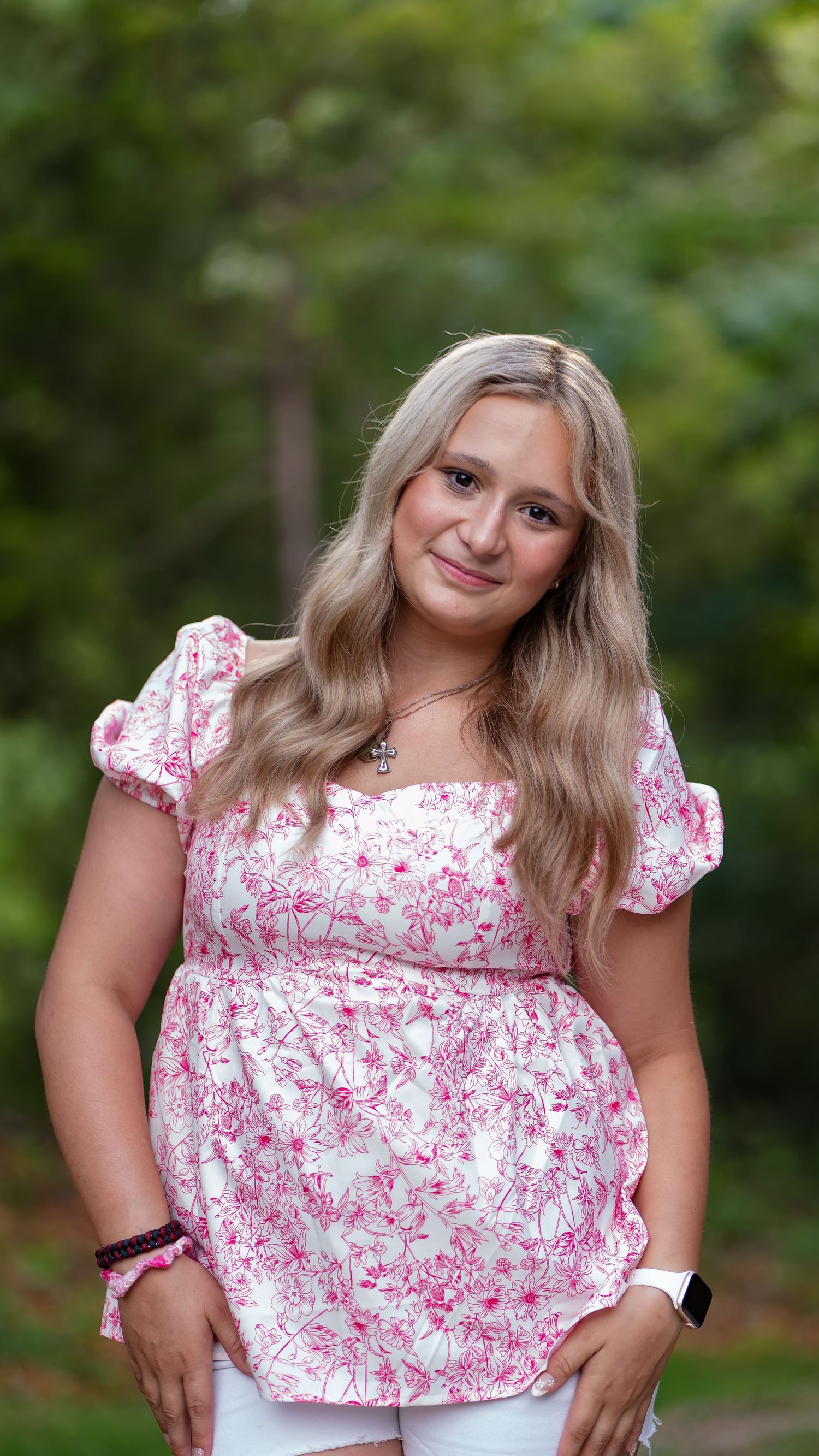A person in a pink and white floral dress poses outdoors against a natural green background.